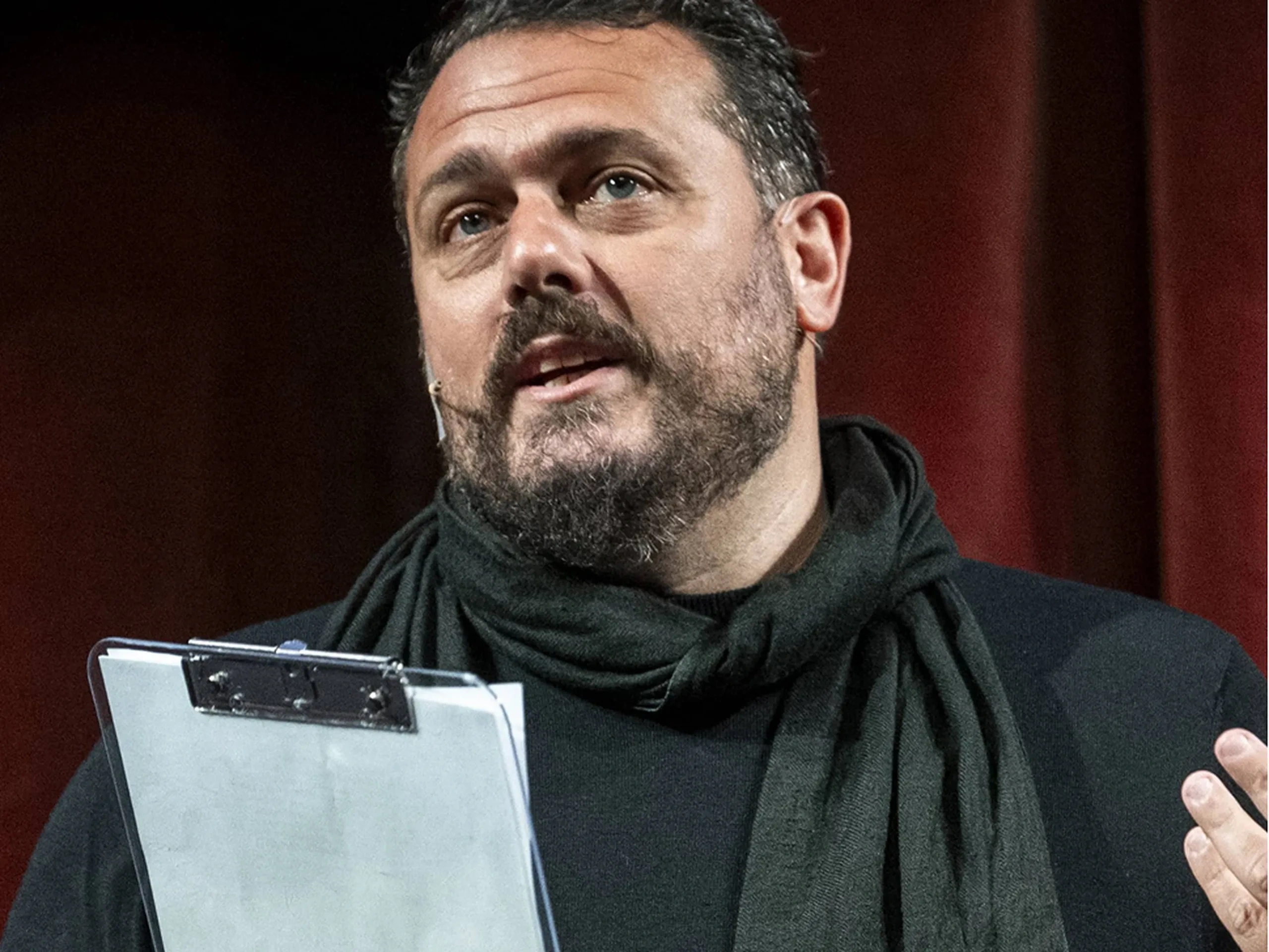 Un uomo barbuto con una sciarpa nera parla con passione sul palco, tenendo in mano un blocco per appunti.