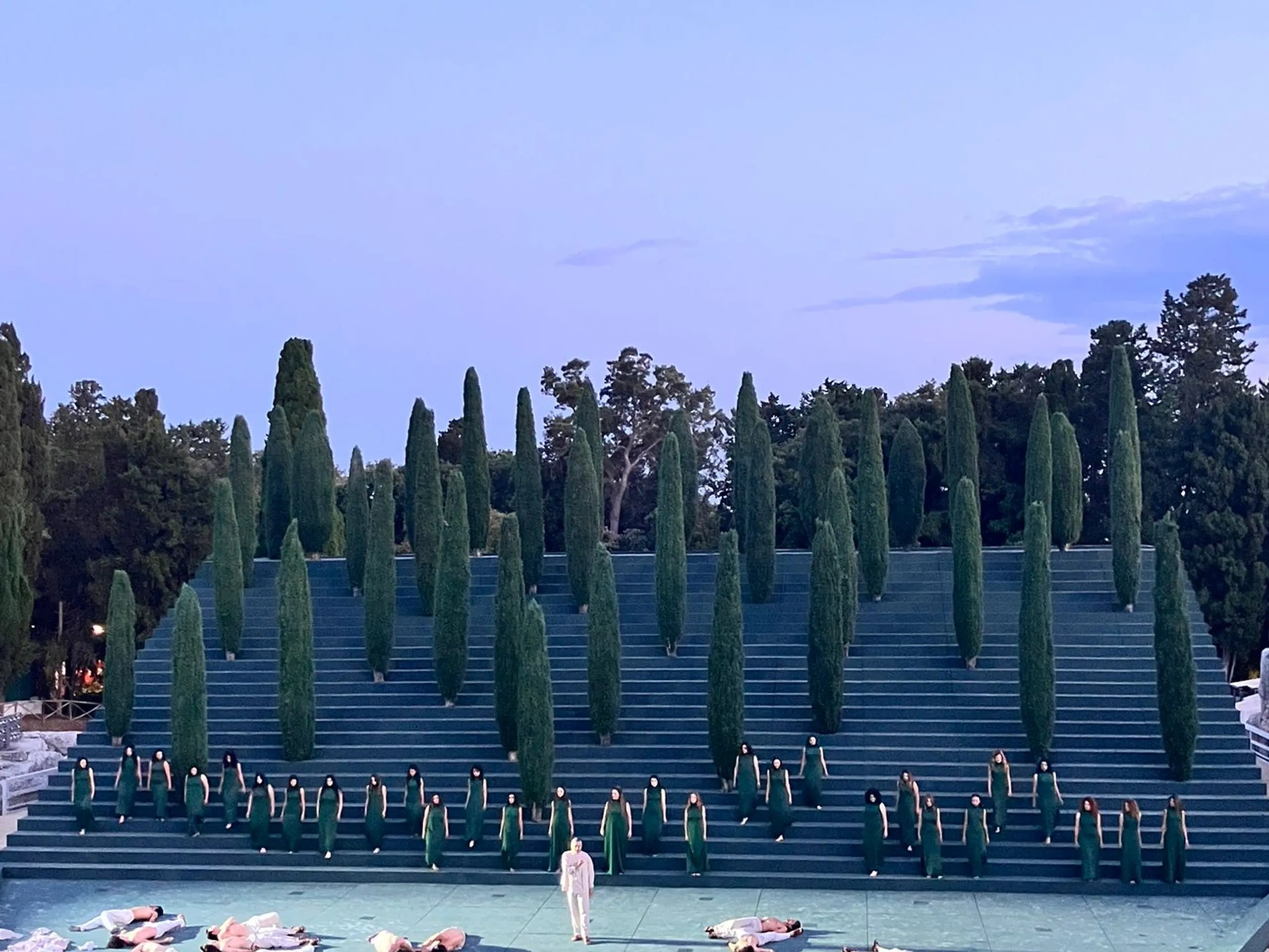 Spettacolo teatrale all'aperto su gradinate con alberi disposti a intervalli regolari; attori vestiti di bianco distesi sul palco, pubblico che assiste.