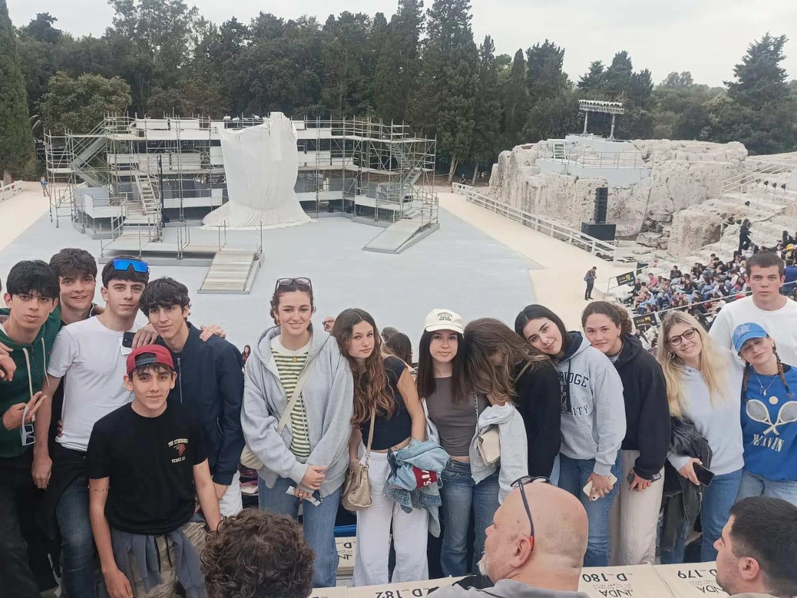 Un gruppo di giovani in posa in un antico teatro all'aperto con gradinate in pietra e un palcoscenico con impalcature e una scultura drappeggiata.