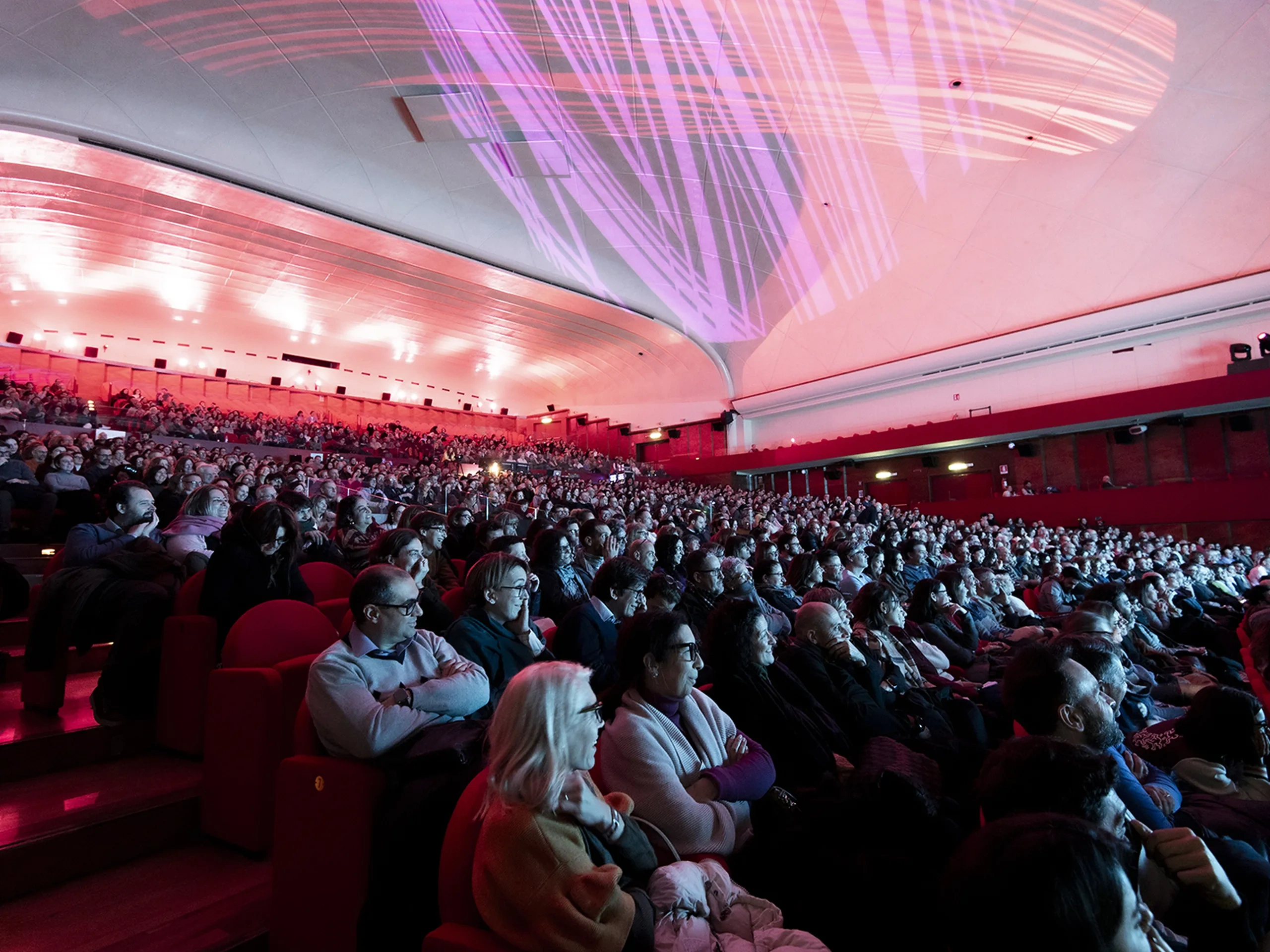 Audience seated in a large theater with a curved ceiling, illuminated by red and purple lights from a stage performance or presentation.