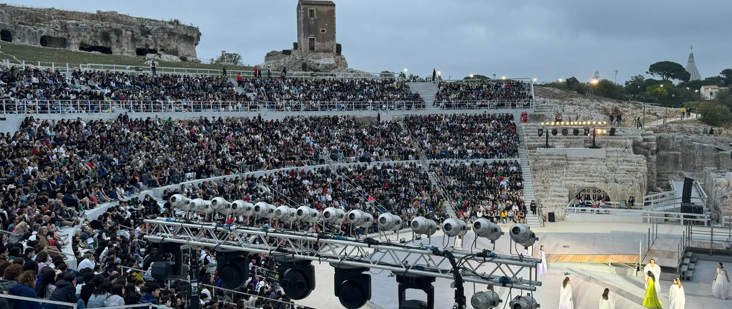 Un grande anfiteatro antico all'aperto gremito di spettatori. Sul palco si esibiscono artisti in costumi colorati. Sullo sfondo sono visibili delle rovine.