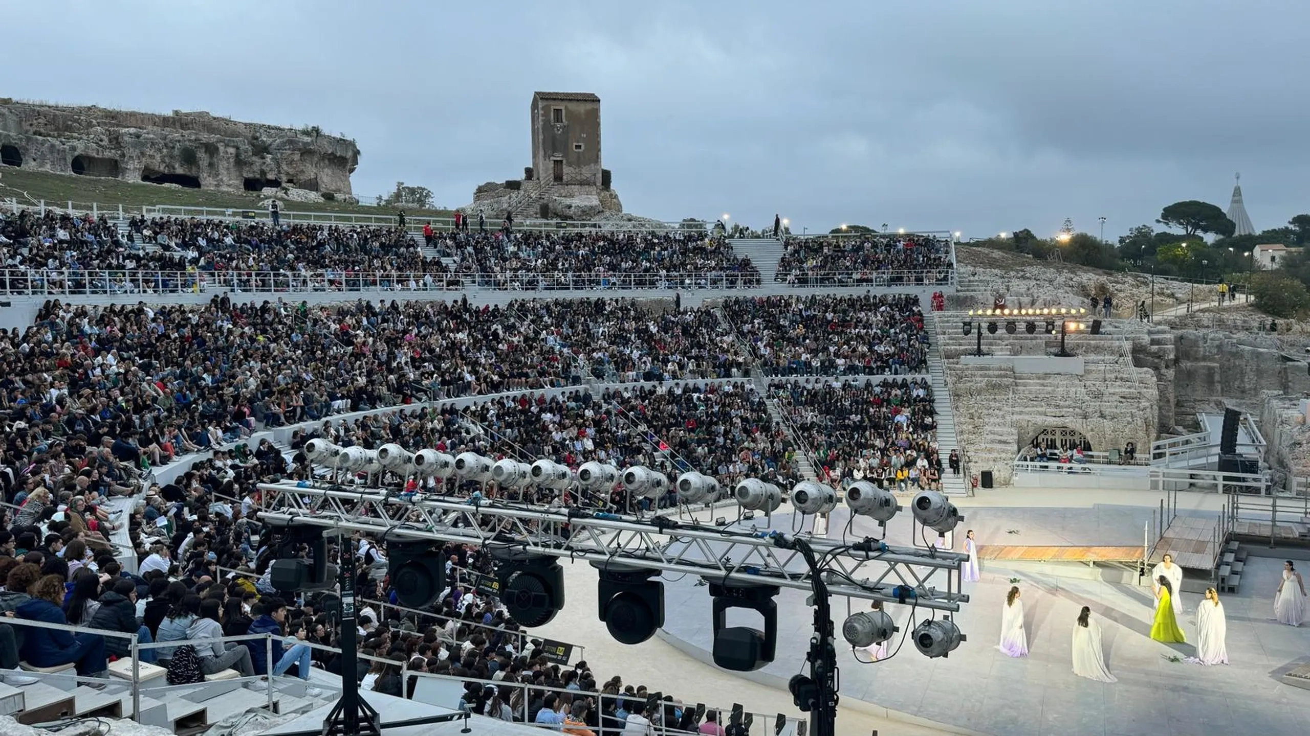 Un grande anfiteatro antico all'aperto gremito di spettatori. Sul palco si esibiscono artisti in costumi colorati. Sullo sfondo sono visibili delle rovine.