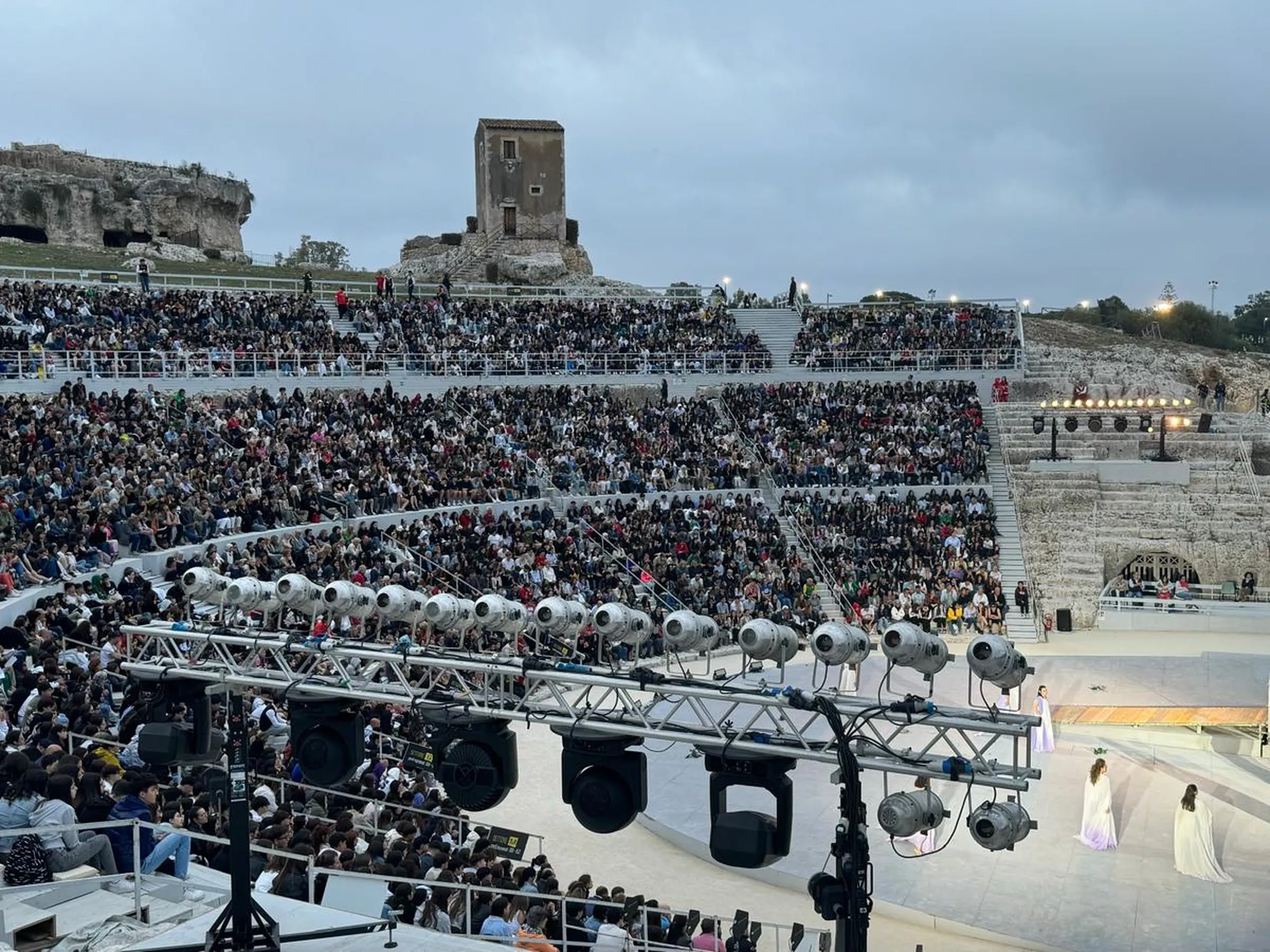 Un grande anfiteatro antico all'aperto gremito di spettatori. Sul palco si esibiscono artisti in costumi colorati. Sullo sfondo sono visibili delle rovine.