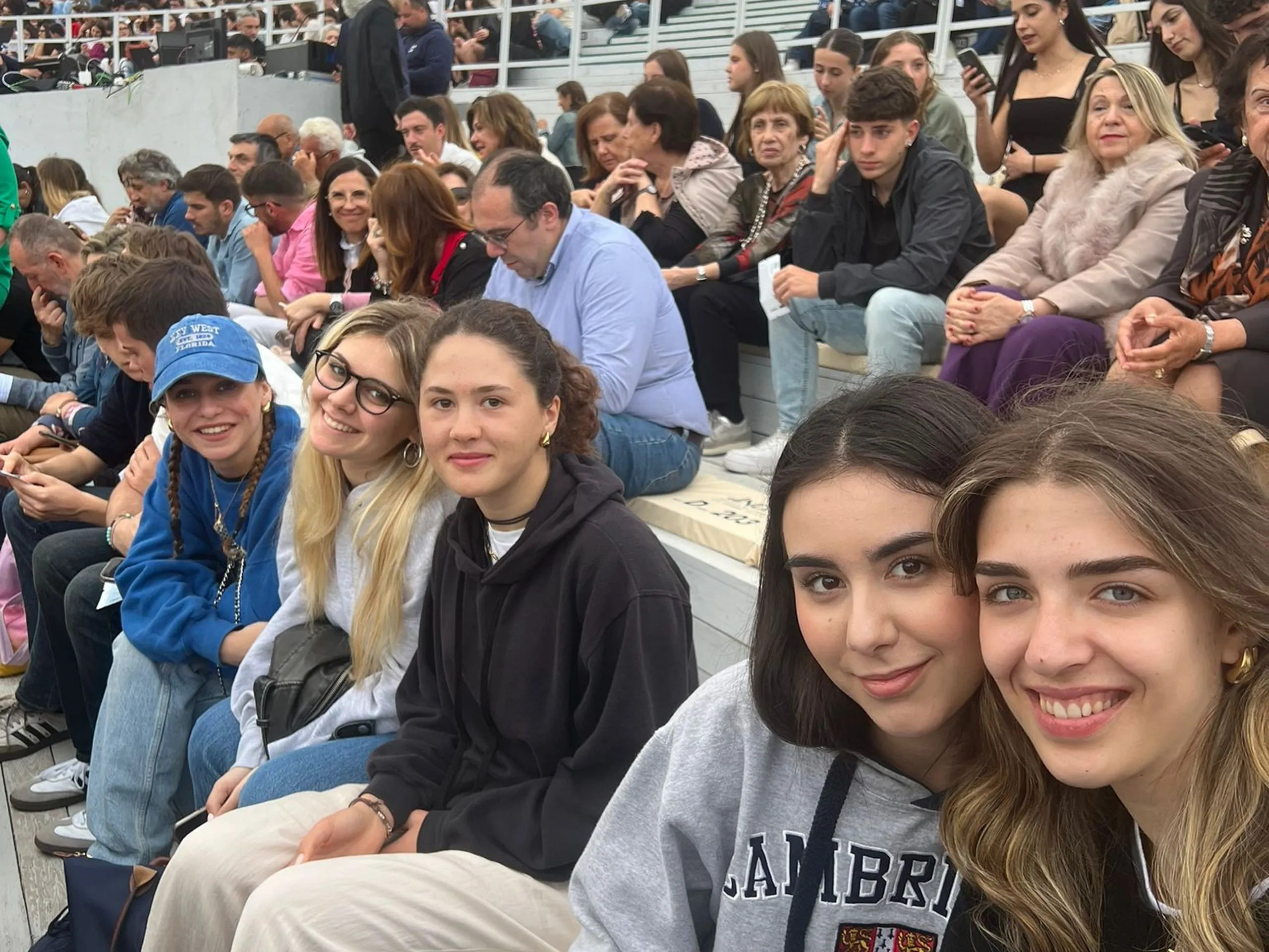 Un gruppo di giovani si scatta un selfie durante un evento all'aperto, con una folla seduta sullo sfondo.
