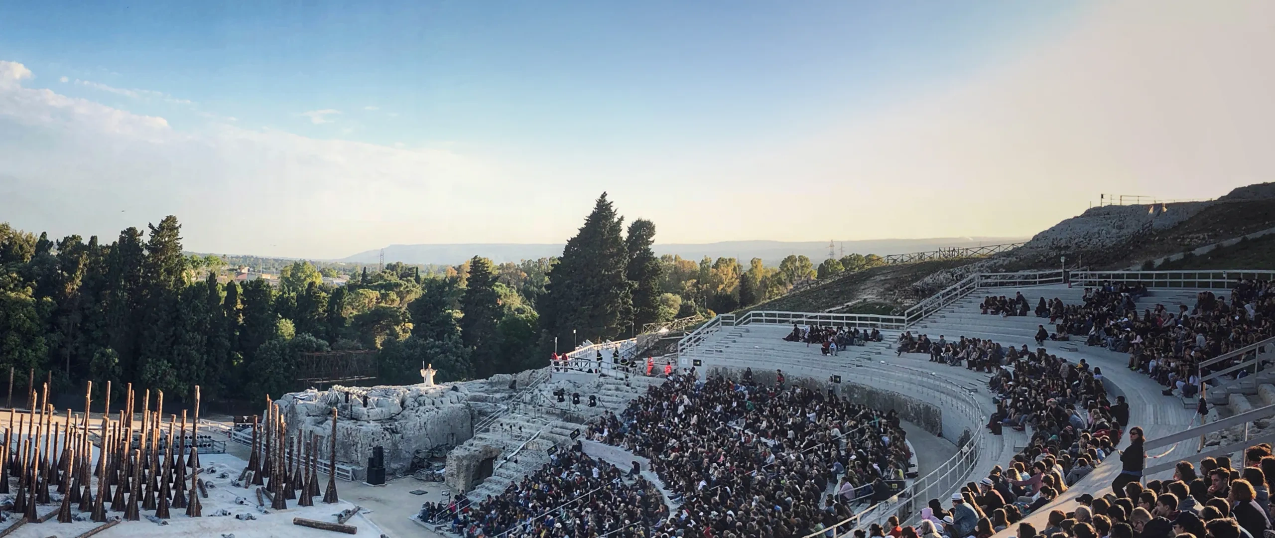 Folla seduta in un antico anfiteatro all'aperto con un palco sulla sinistra, circondato da alberi e una vista lontana del paesaggio sotto un cielo limpido.