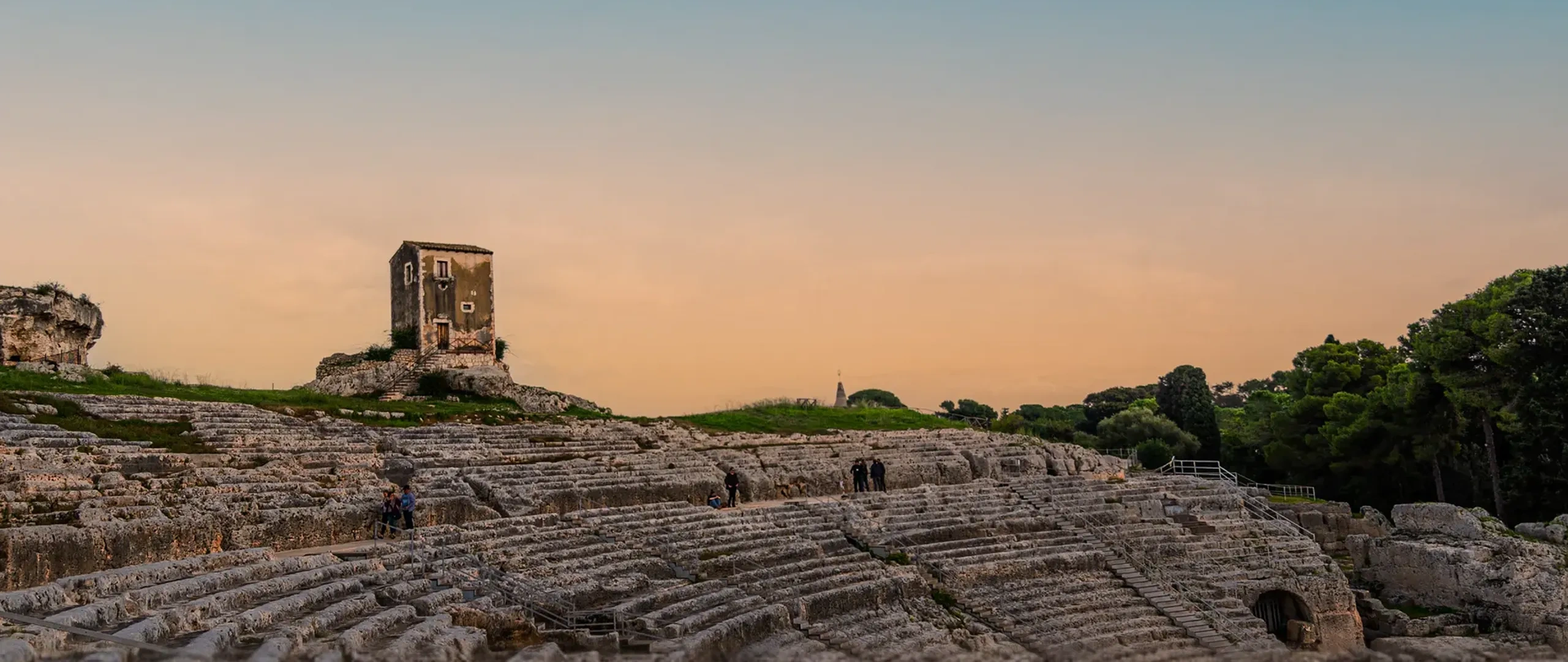 Antico anfiteatro in pietra con visitatori sparsi qua e là, circondato da alberi e da una torre rettangolare solitaria sotto un cielo azzurro e limpido al tramonto.