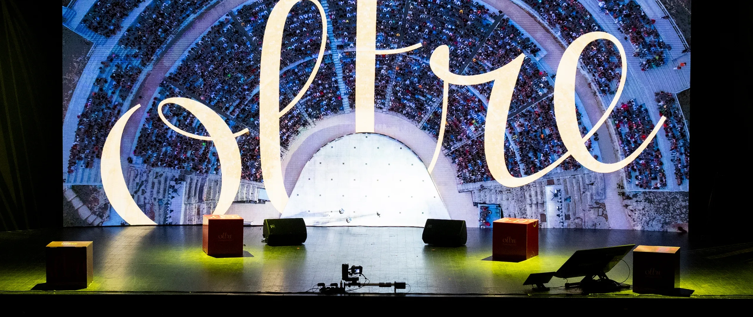 Grande palco con uno schermo che mostra la scritta “oltre” su una veduta aerea di una folla. Sul palco ci sono quattro cubi e delle attrezzature.
