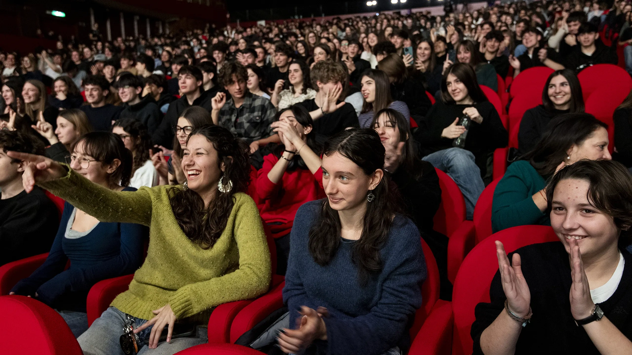 Un folto pubblico seduto in un teatro, sorridente e applaudente. Molti indossano abiti casual e le poltrone rosse creano un'atmosfera vivace