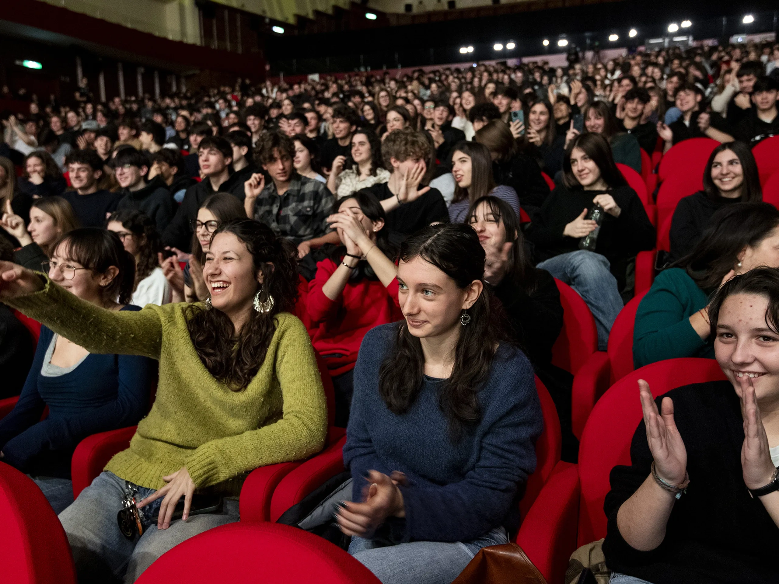 Un folto pubblico seduto in un teatro, sorridente e applaudente. Molti indossano abiti casual e le poltrone rosse creano un'atmosfera vivace
