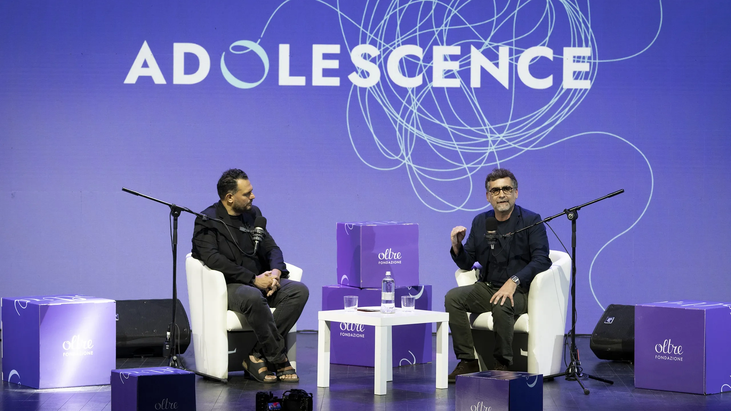 Due uomini seduti sul palco con i microfoni, intenti a discutere sotto un grande cartello con la scritta «ADOLESCENCE», circondati da scatole viola della Oltre Fondazione.