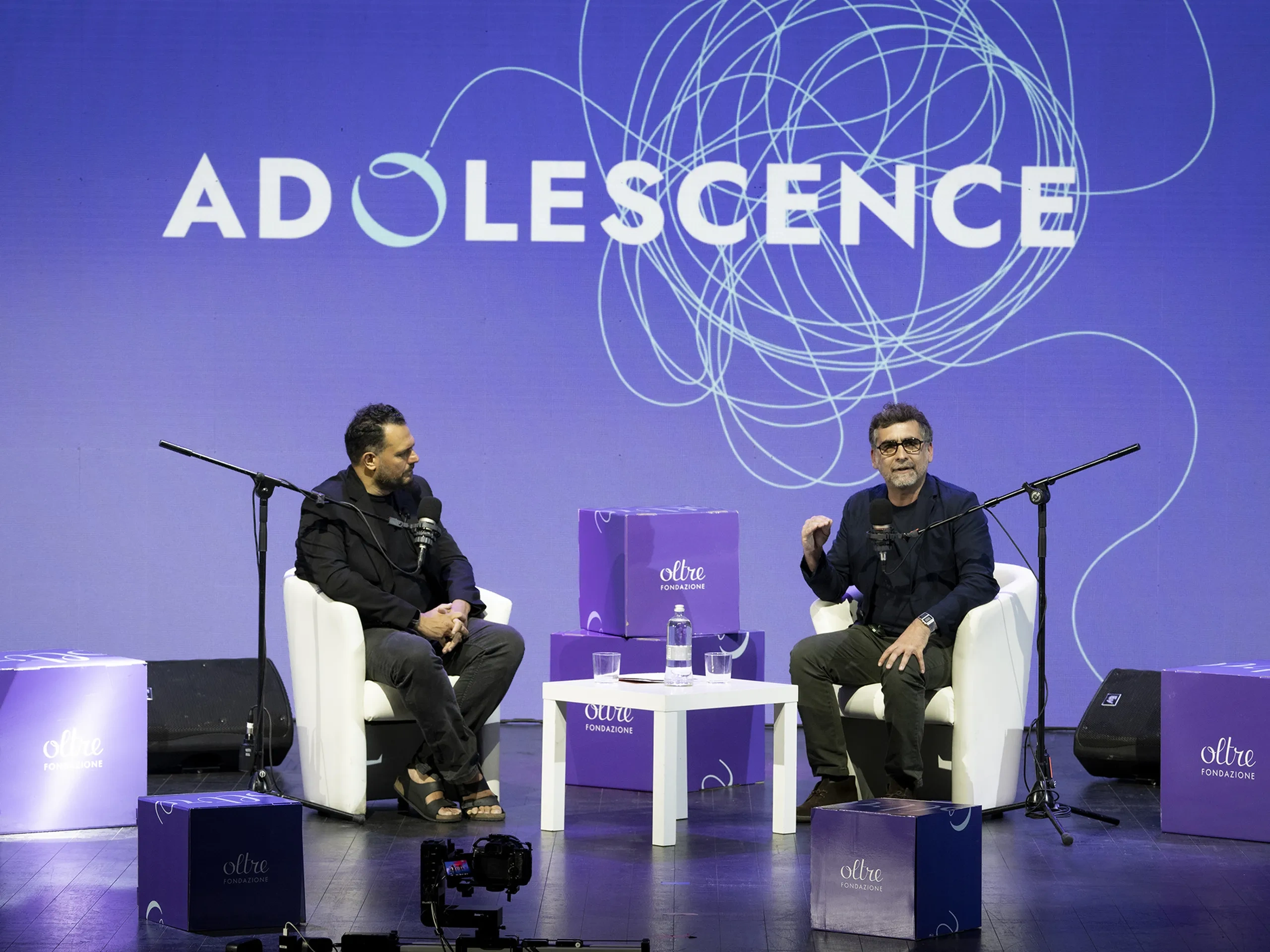 Due uomini seduti sul palco con i microfoni, intenti a discutere sotto un grande cartello con la scritta «ADOLESCENCE», circondati da scatole viola della Oltre Fondazione.