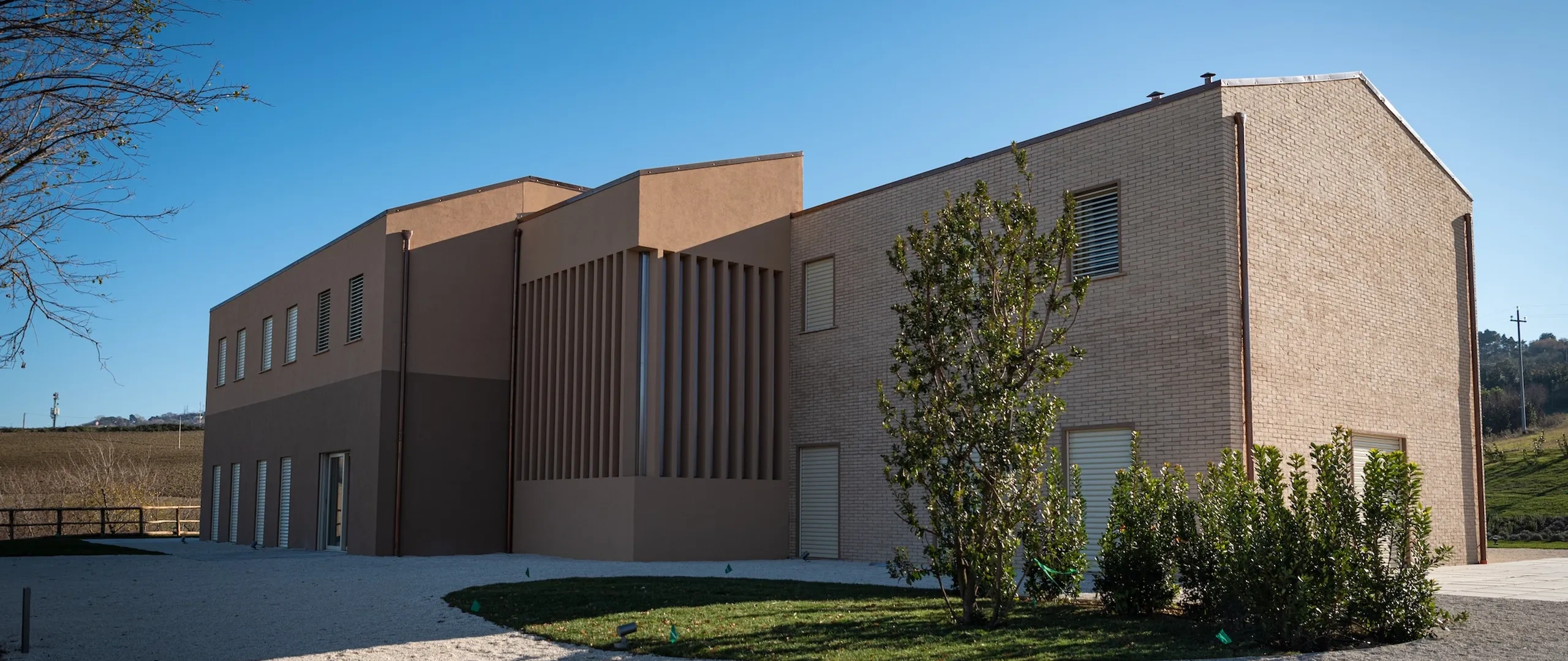 Modern two-story building with a mix of brown and tan brickwork, surrounded by a gravel path and greenery under a clear blue sky.