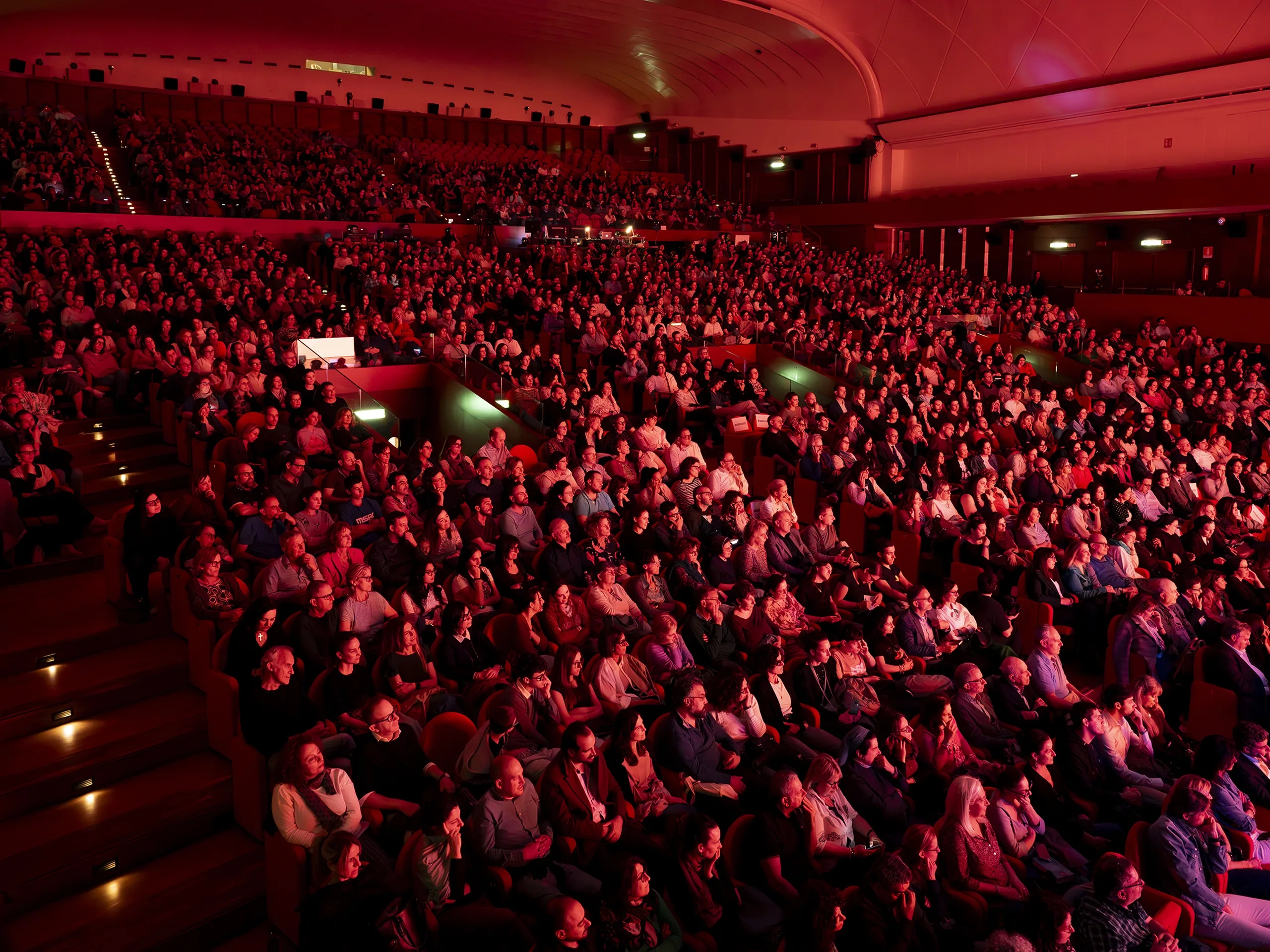 A large audience seated in a dimly lit theater with red lighting, attentively watching a performance or event.