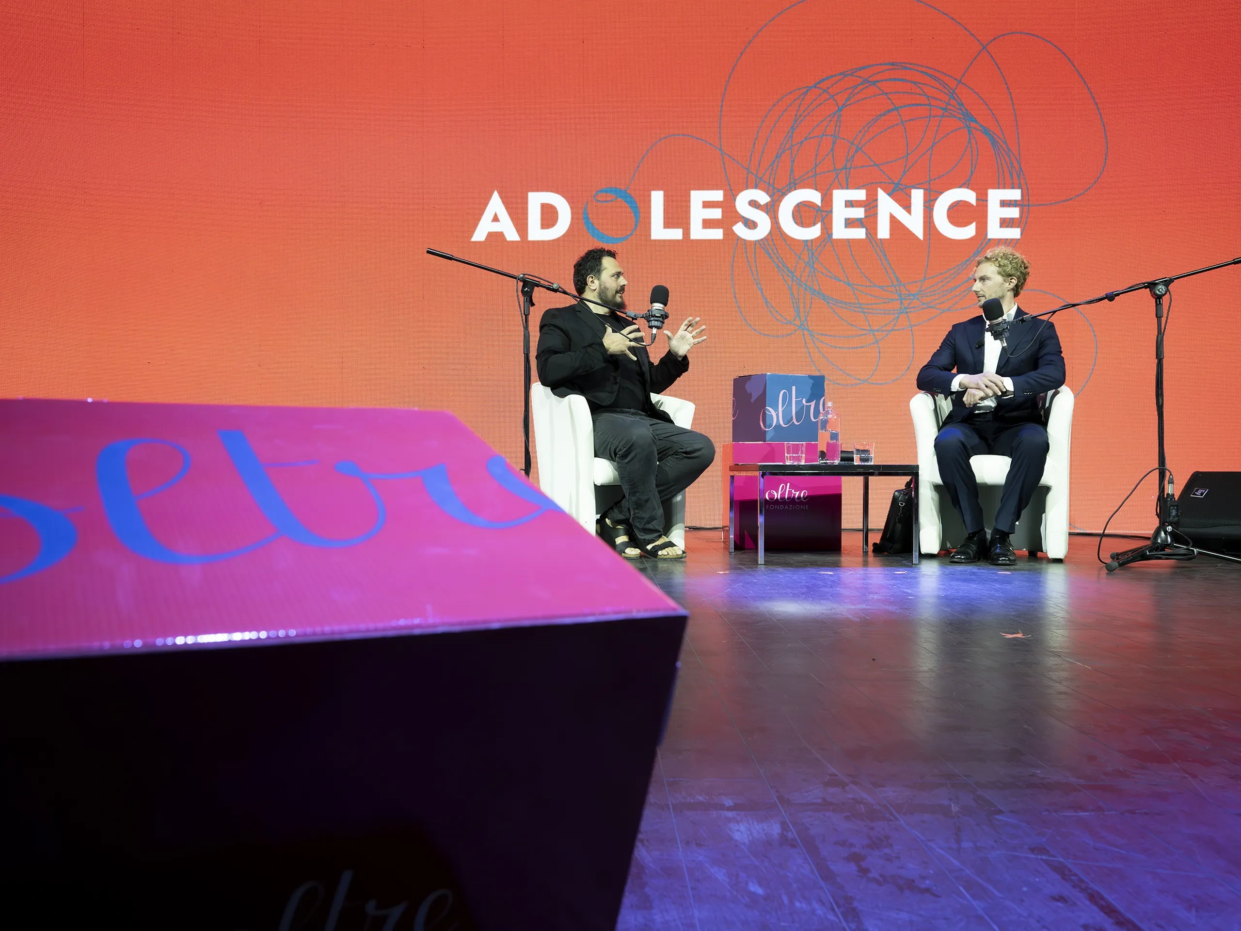Two people seated on stage discussing adolescence, with microphones and a backdrop displaying the word "ADOLESCENCE" in bold letters.