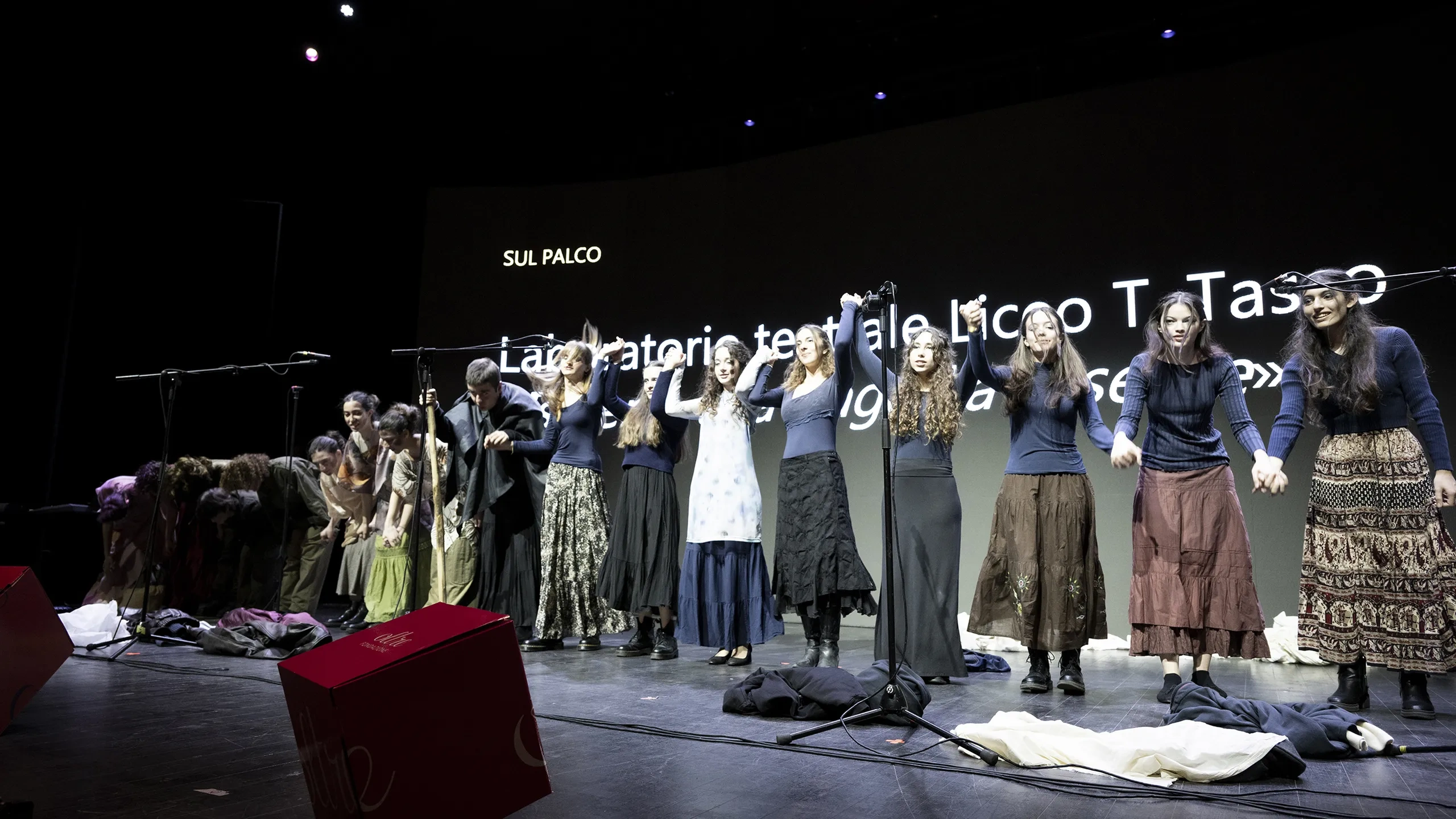 Gli artisti salutano il pubblico sul palco tenendosi per mano, con uno sfondo proiettato su cui è visualizzato del testo.