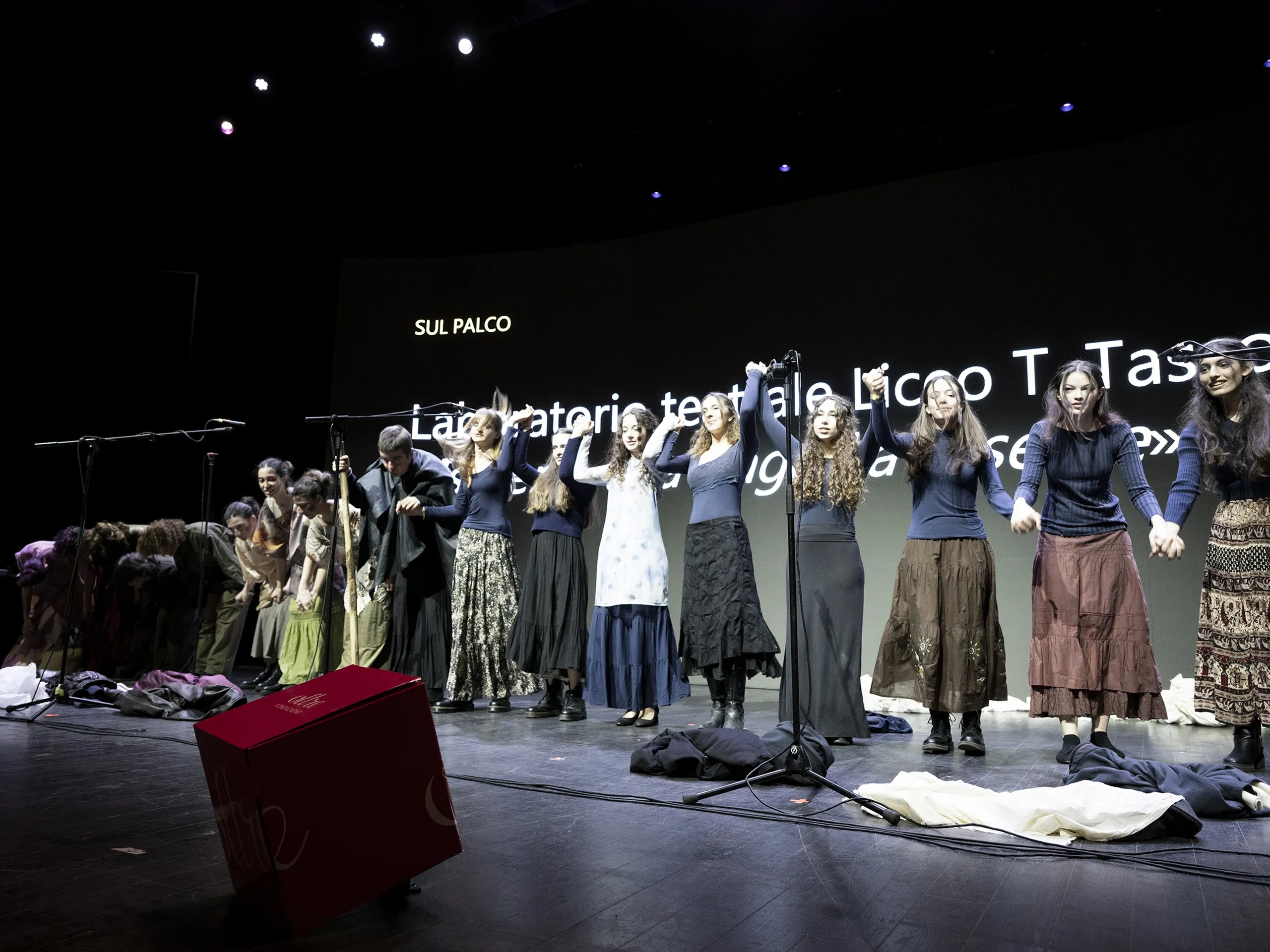 Gli artisti salutano il pubblico sul palco tenendosi per mano, con uno sfondo proiettato su cui è visualizzato del testo.