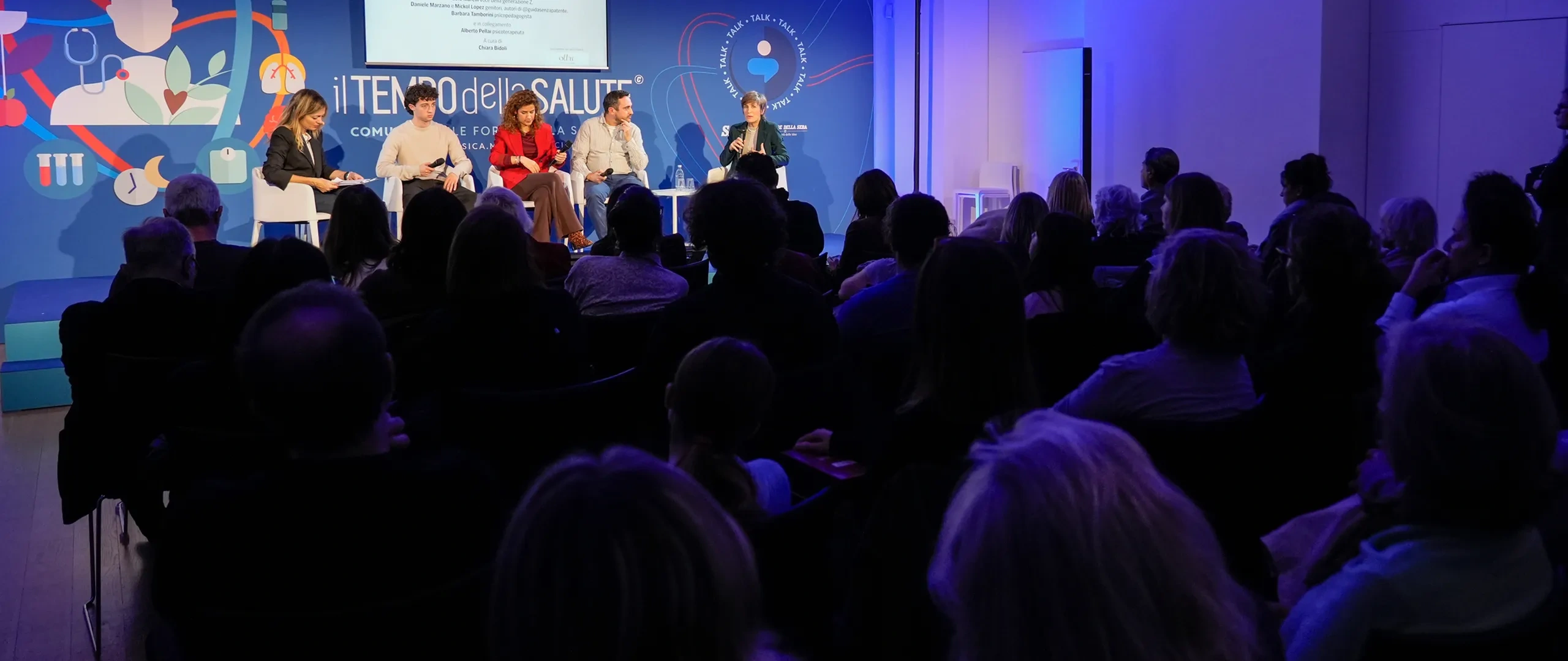 Il pubblico in silhouette assiste a una tavola rotonda su un palco illuminato di blu con cinque relatori seduti, uno schermo per le presentazioni e uno sfondo a tema salute.