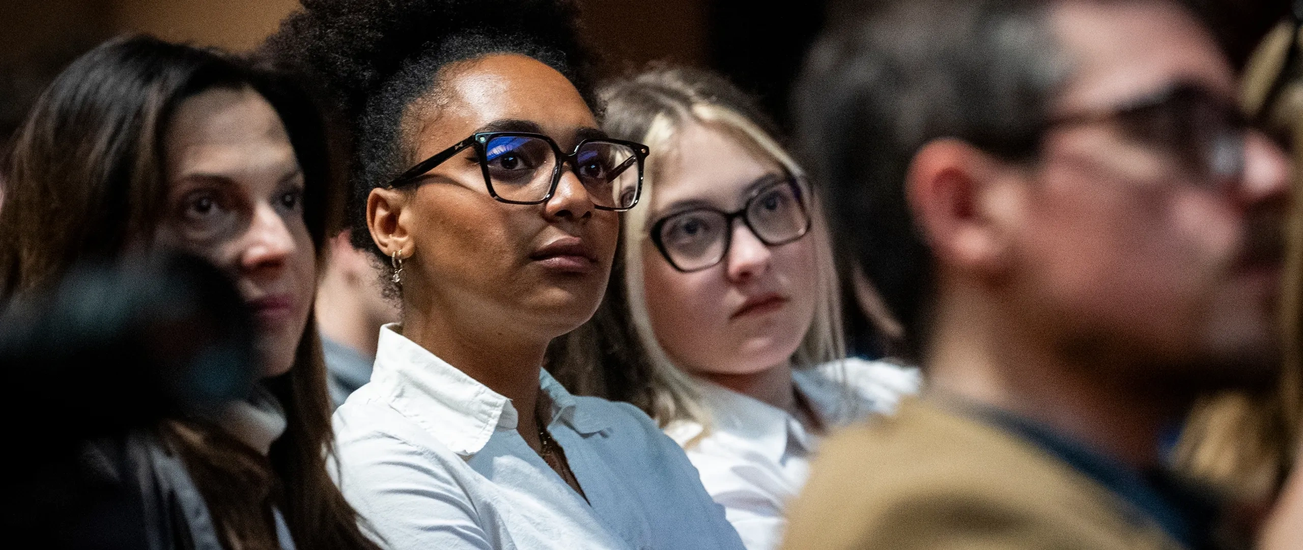 A group of attentive people seated indoors, listening. The focus is on a woman with glasses and curly hair, wearing a white shirt.