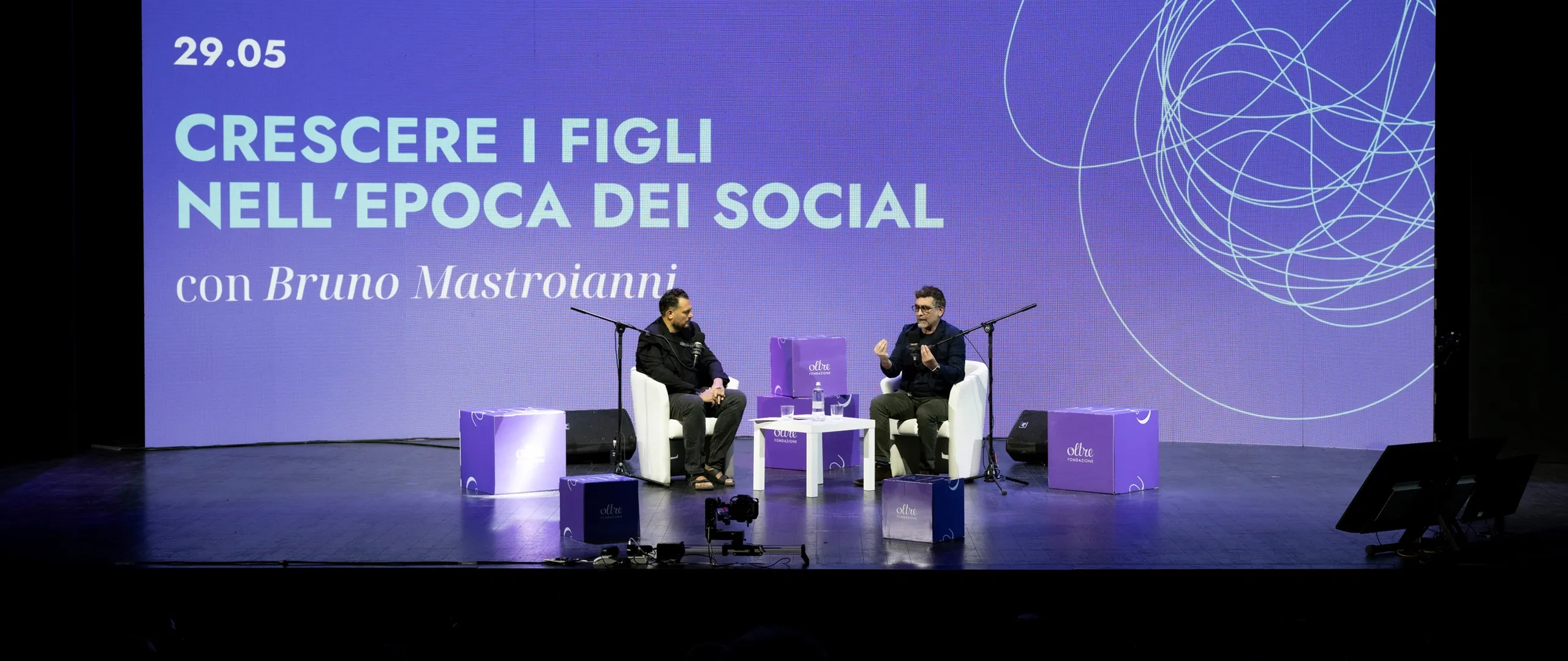 Due relatori sul palco discutono del tema «Crescere i figli nell'era dei social media» su uno sfondo di linee vorticose e testo in italiano.