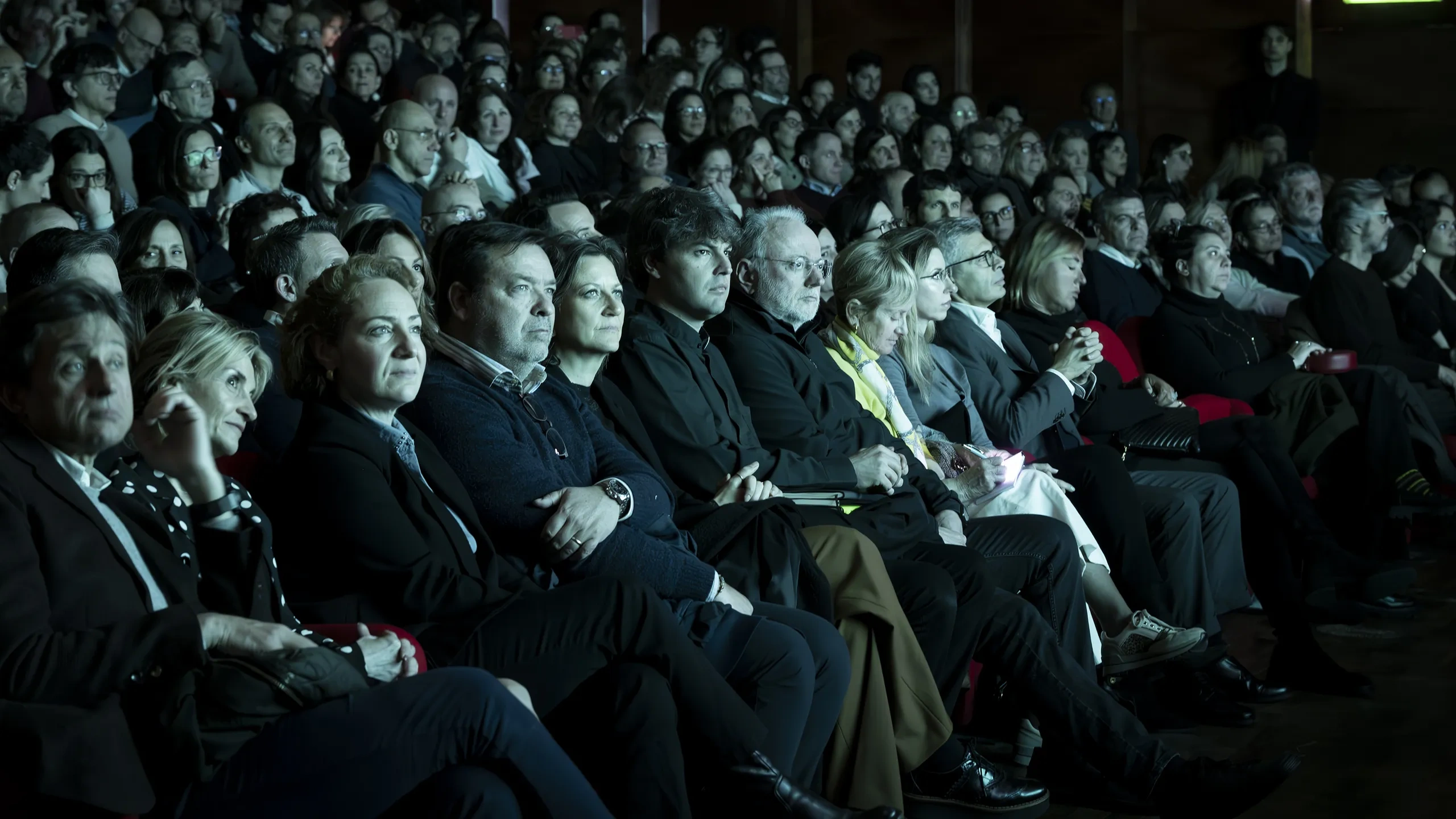Un folto pubblico è seduto in silenzio in un teatro in penombra; la maggior parte delle persone è rivolta verso il palcoscenico, mentre alcune sono illuminate da una luce soffusa.