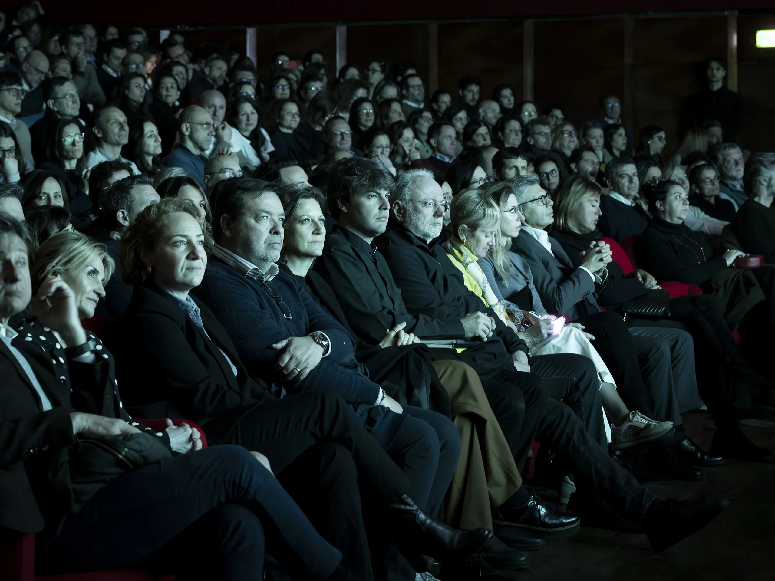 Un folto pubblico è seduto in silenzio in un teatro in penombra; la maggior parte delle persone è rivolta verso il palcoscenico, mentre alcune sono illuminate da una luce soffusa.