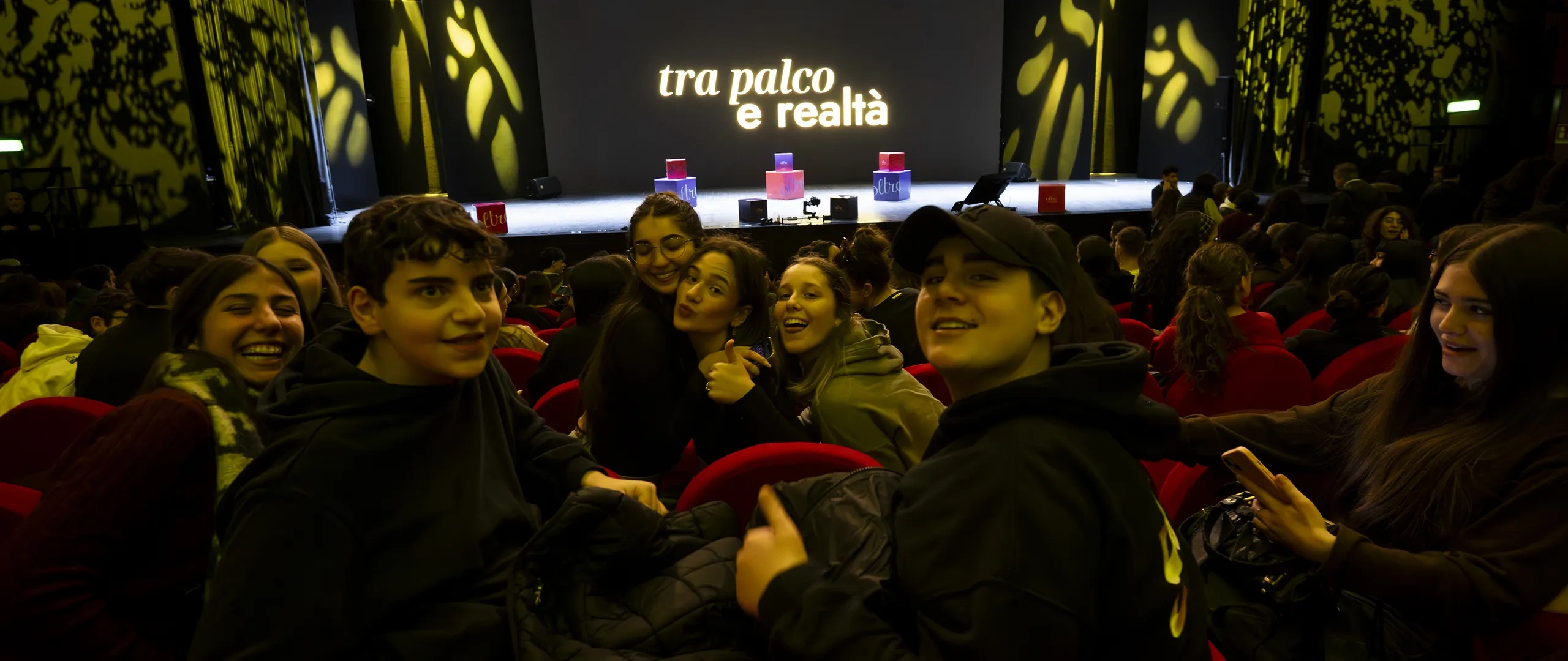 Audience members seated in a theater, facing a stage with two chairs and a backdrop displaying "tra palco e realtà."