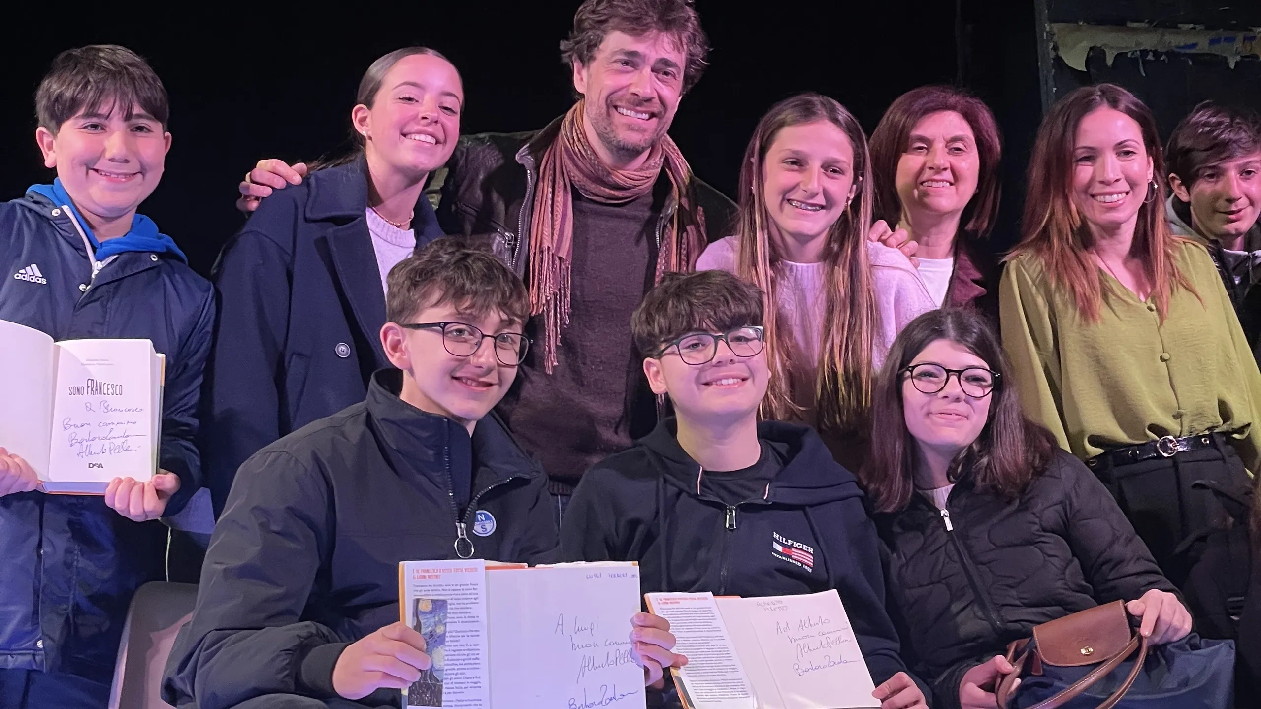Un gruppo di persone sorridenti posa insieme su un palco; due persone in primo piano tengono in mano dei libri aperti con delle firme ben visibili.