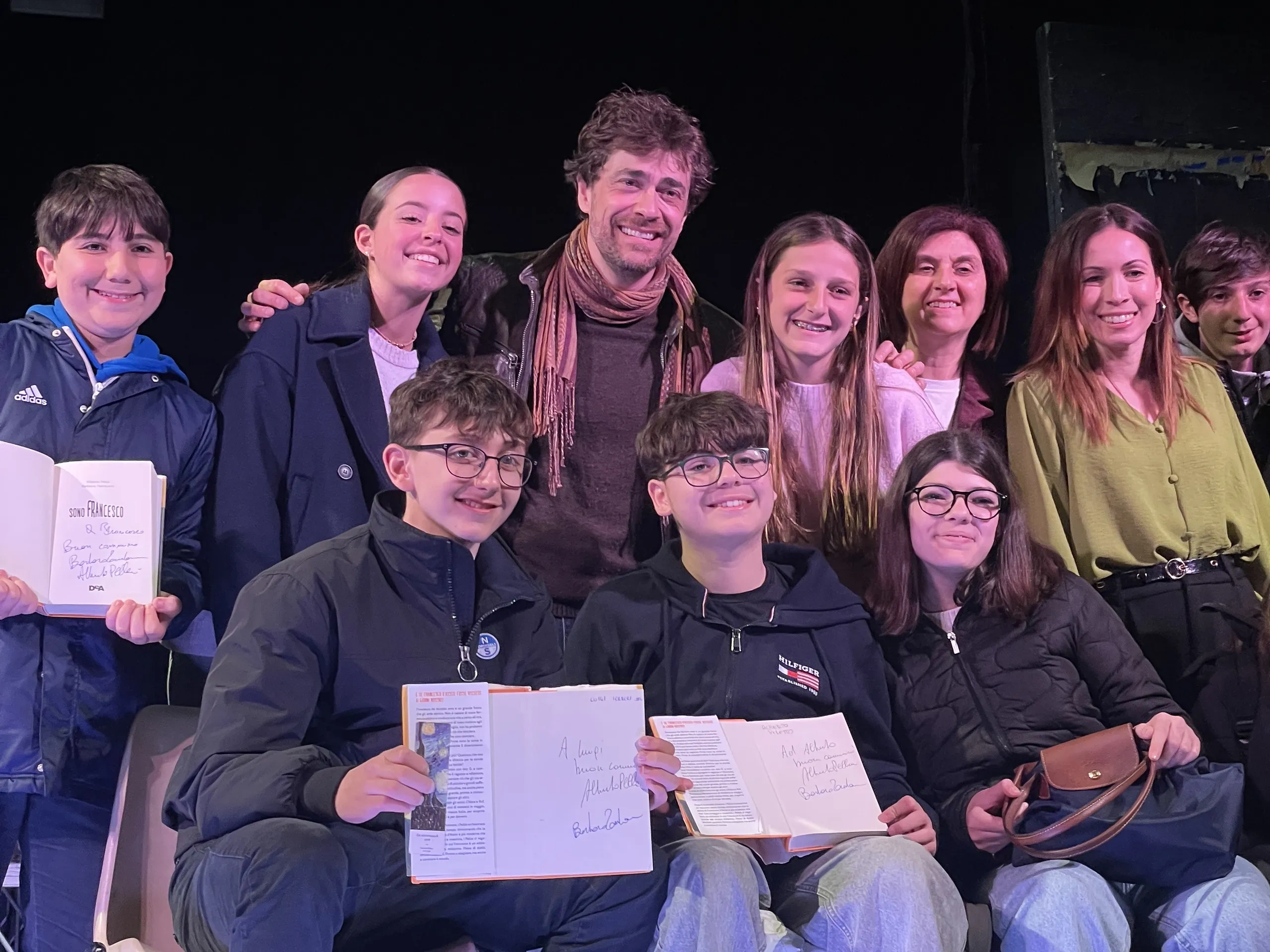 Un gruppo di persone sorridenti posa insieme su un palco; due persone in primo piano tengono in mano dei libri aperti con delle firme ben visibili.