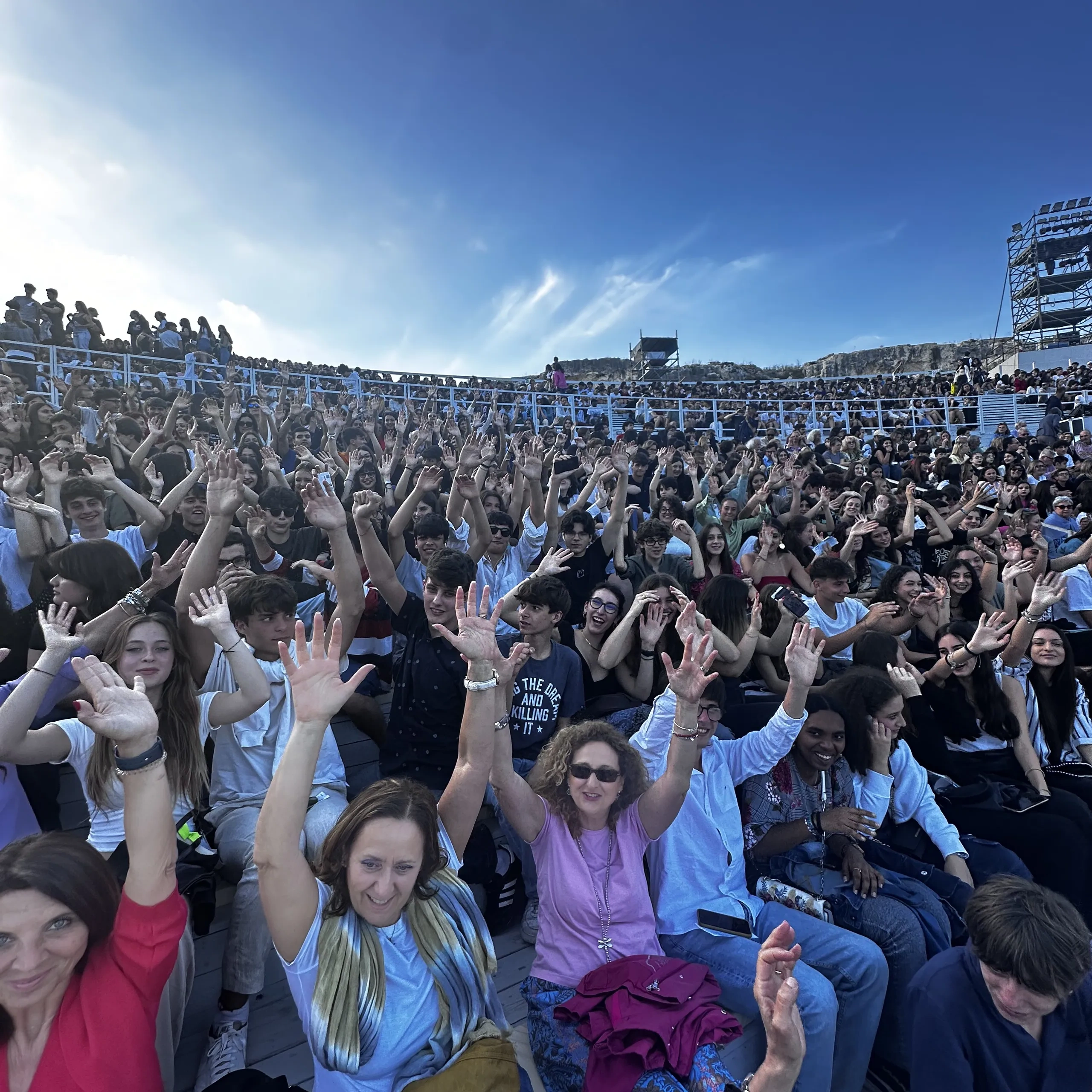 Una grande folla di persone sedute in uno stadio, molte delle quali con le mani alzate sotto un cielo azzurro e limpido.
