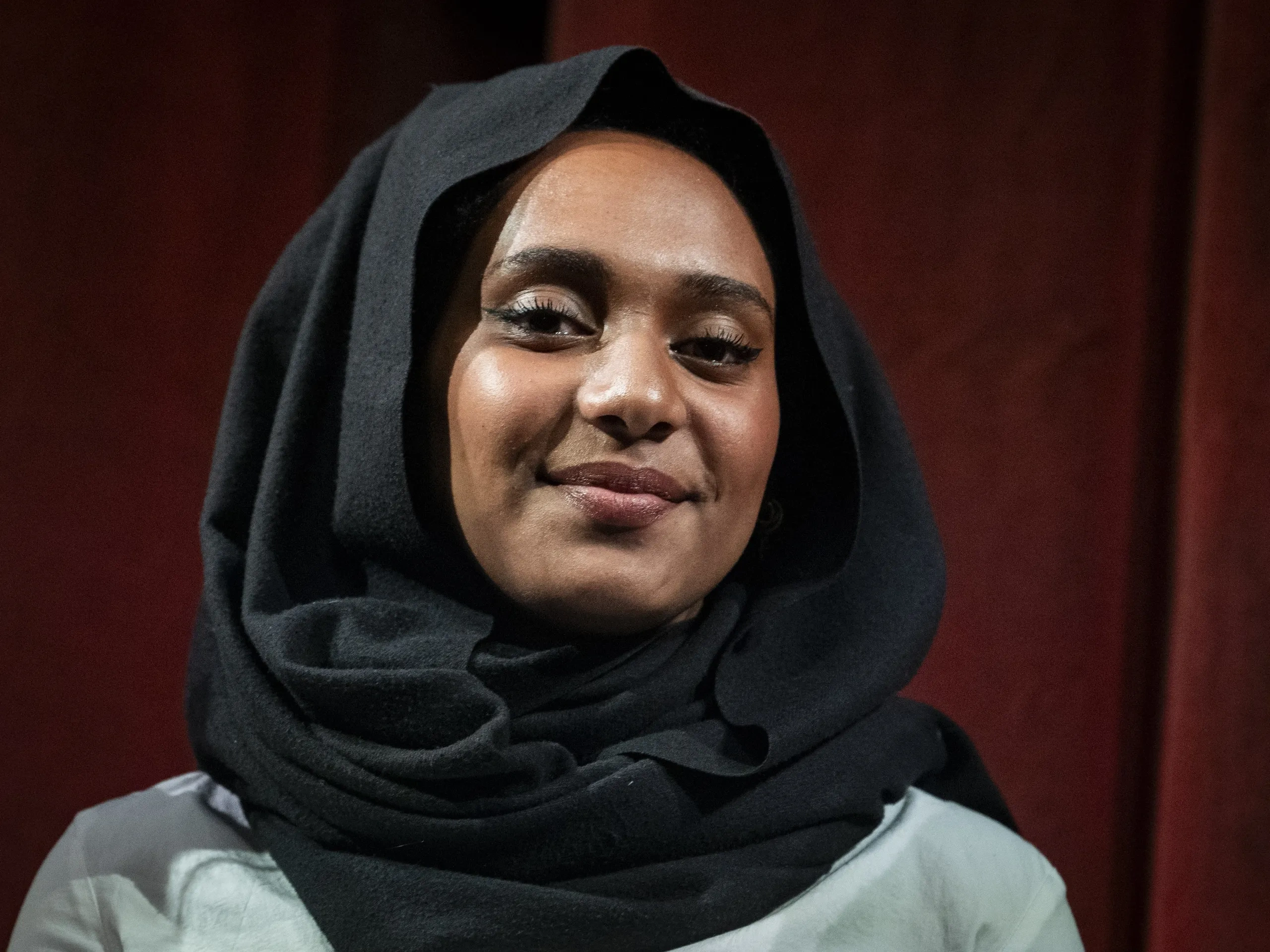 Una persona sorridente, che indossa un hijab nero e una camicia bianca, è in piedi su uno sfondo rosso scuro.