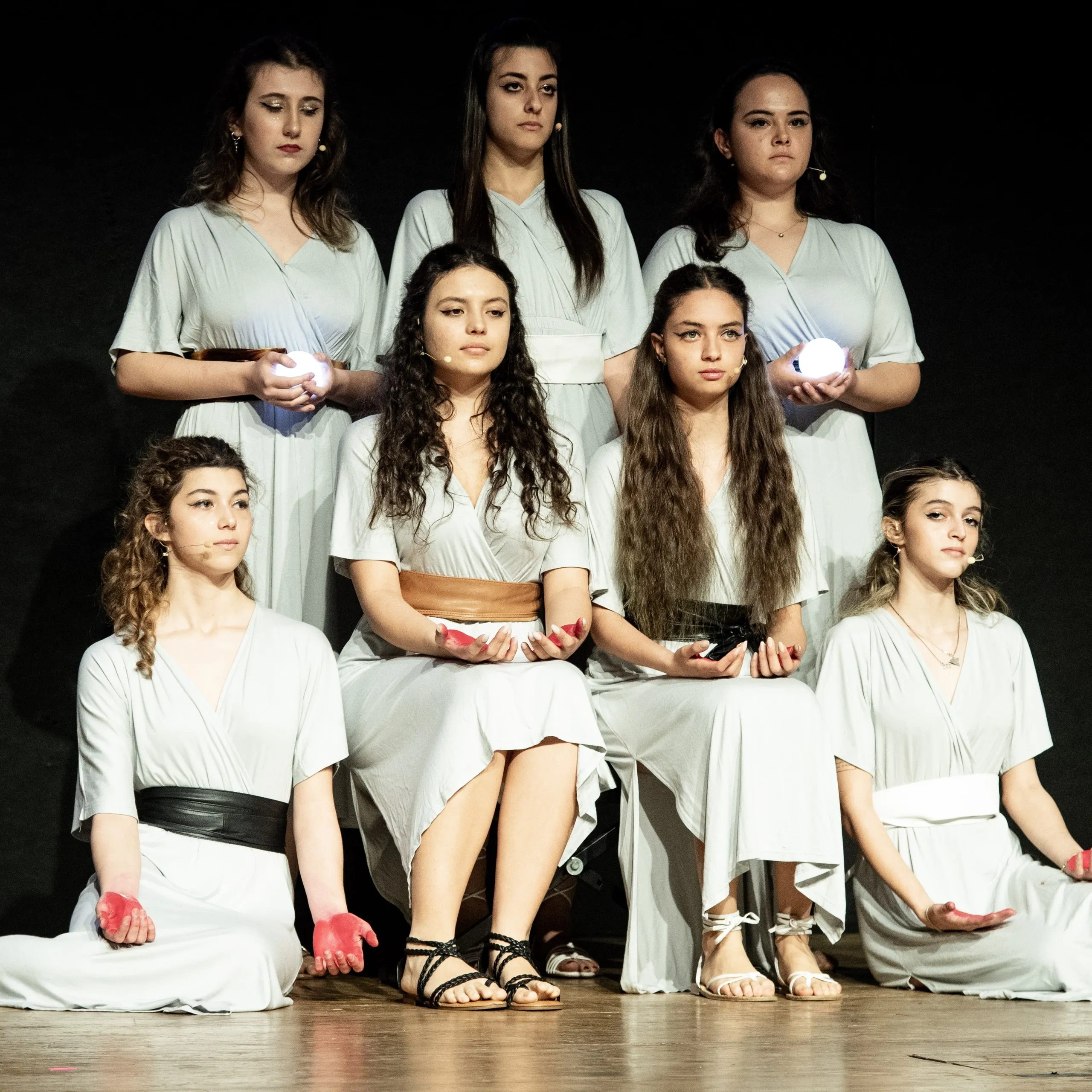 Sette donne in abiti bianchi si esibiscono sul palco, tenendosi per mano con mani luminose o dipinte, in piedi e sedute in una formazione piramidale.