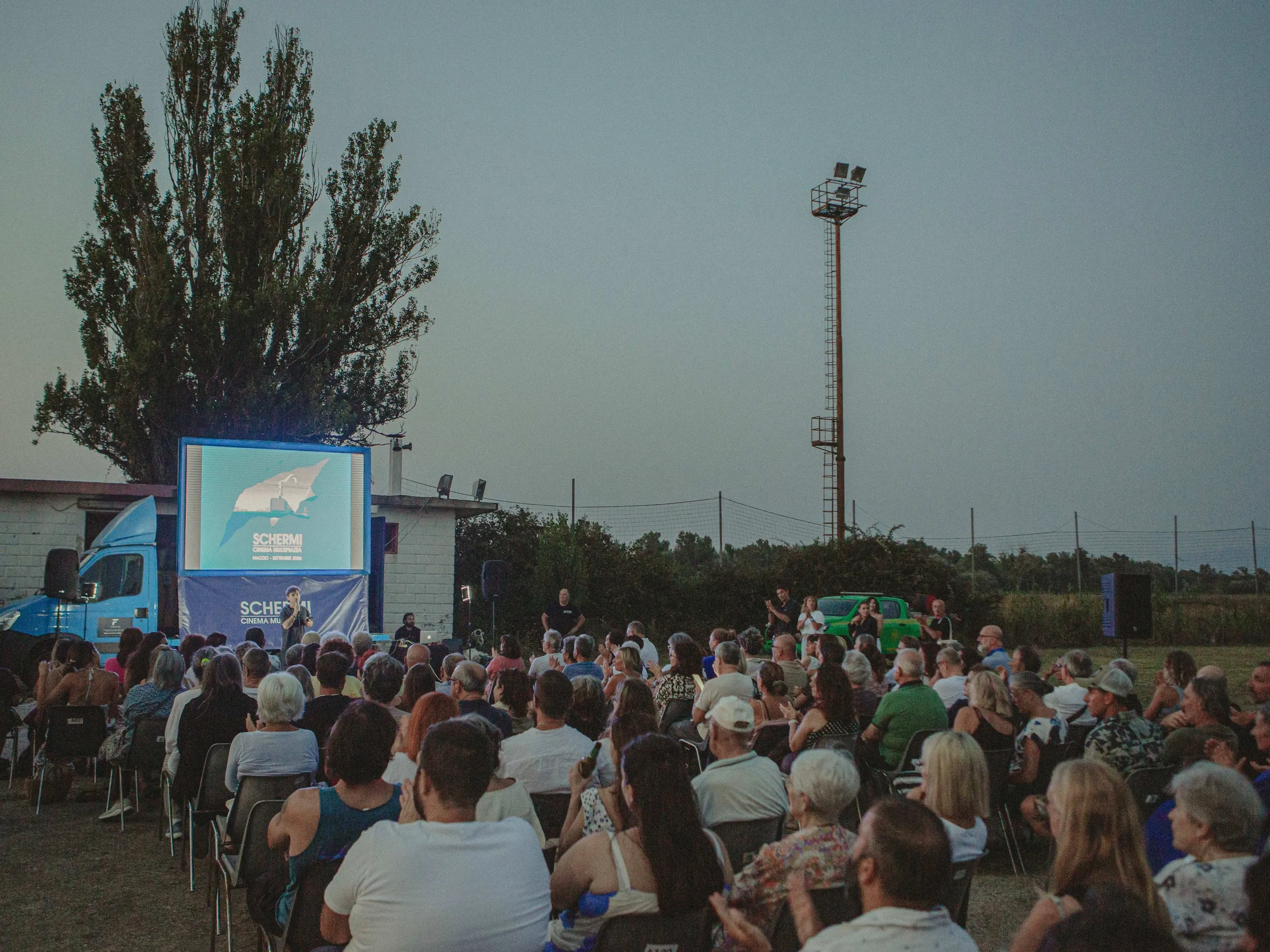 Proiezione cinematografica all'aperto con il pubblico seduto davanti a un grande schermo al crepuscolo, circondato da alberi e dal cielo aperto.