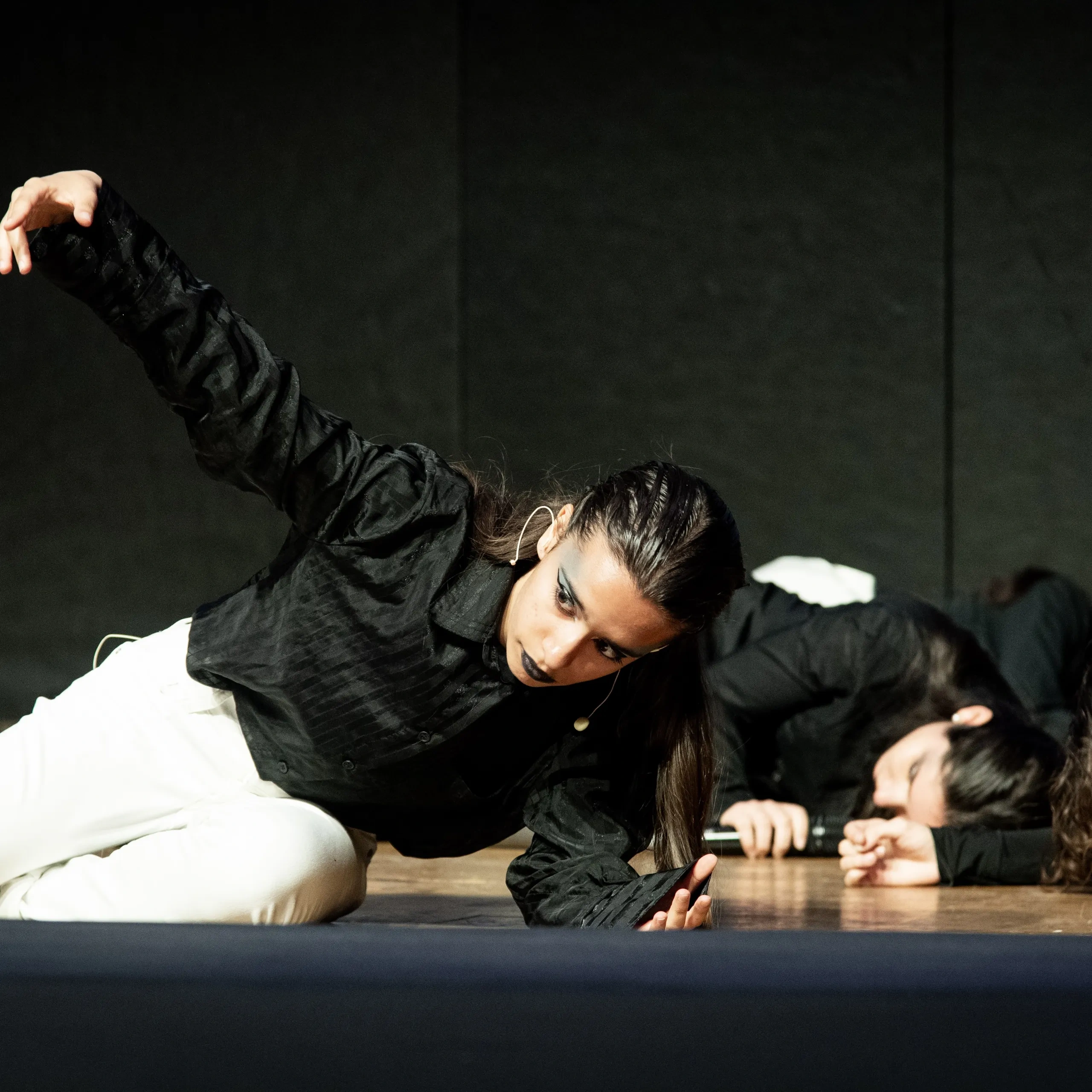 Tre artisti in abiti bianchi e neri distesi su un palco, uno con un braccio disteso, in una posa di danza espressiva.