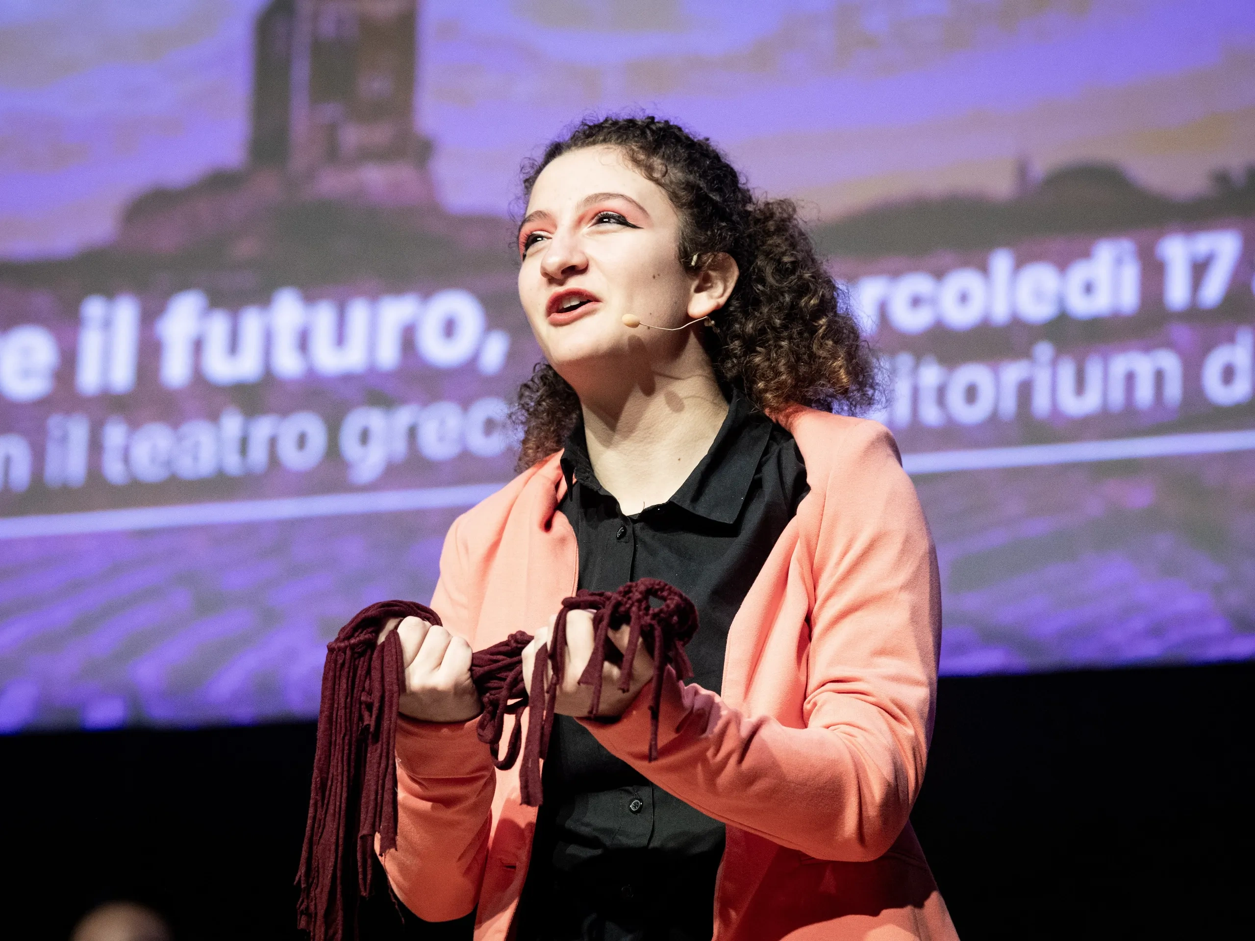 Una persona che parla con passione sul palco, tenendo in mano un oggetto, con uno schermo di presentazione sfocato sullo sfondo.