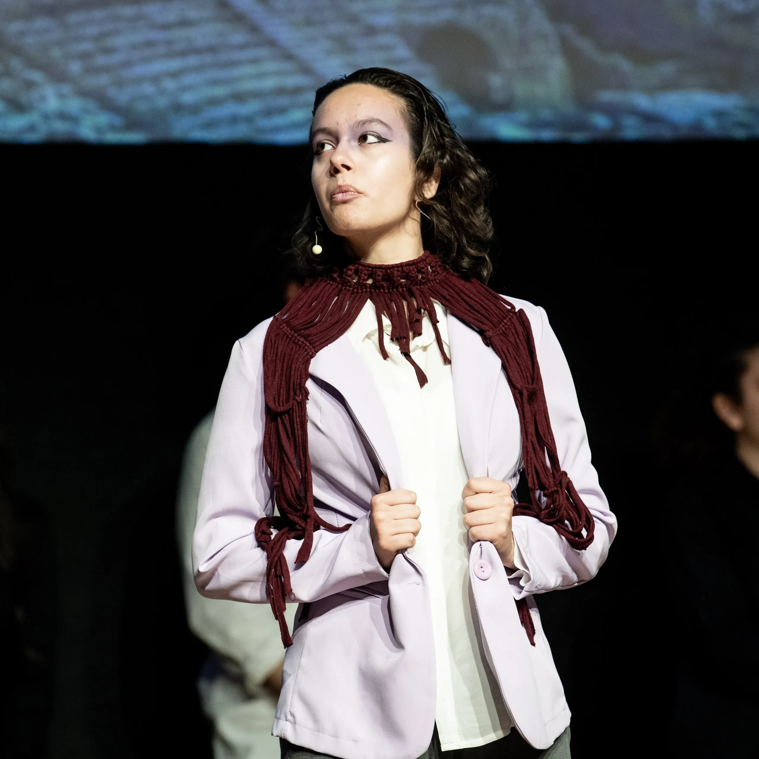 Una donna con un blazer bianco e una sciarpa rossa sta in piedi con sicurezza sul palco, con tre persone sfocate sullo sfondo.
