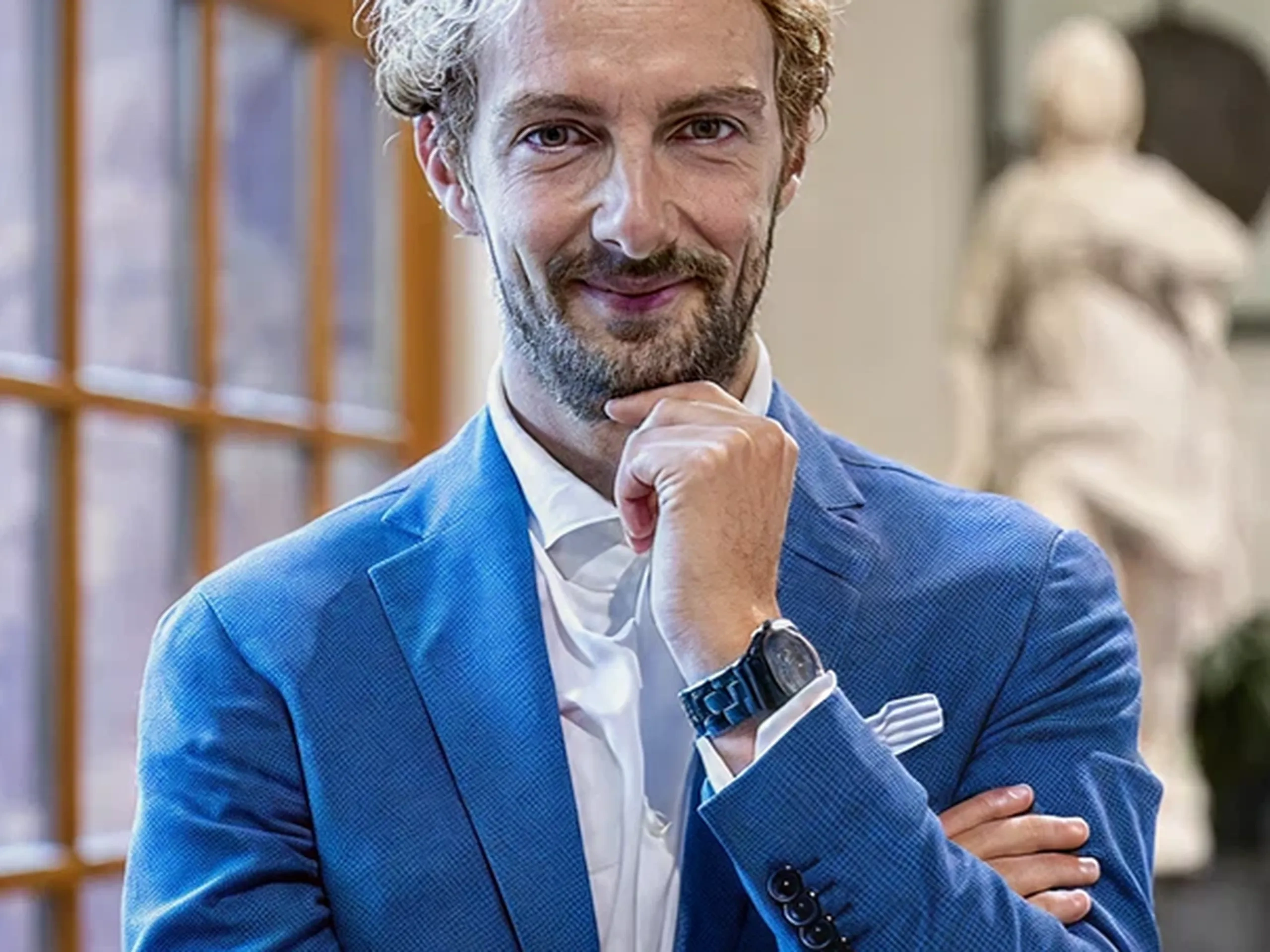 Man with curly hair in blue suit, white shirt, and watch, smiling with hand on chin, standing indoors near a large window.