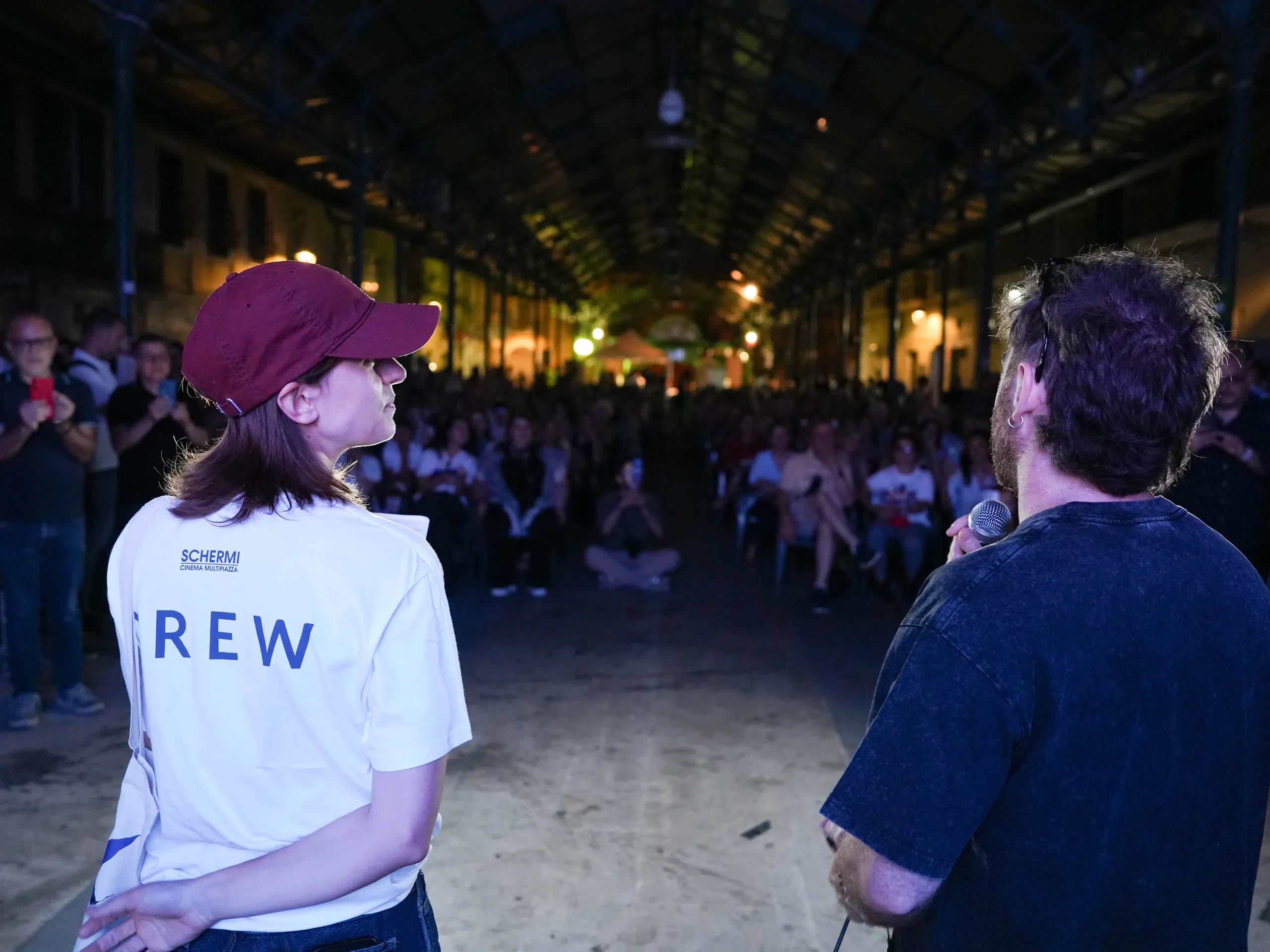Due persone sono in piedi sul palco, di fronte al pubblico, in uno spazio semi-aperto durante la notte. Una tiene in mano un microfono; l'altra indossa una maglietta con la scritta "Crew" e un berretto.