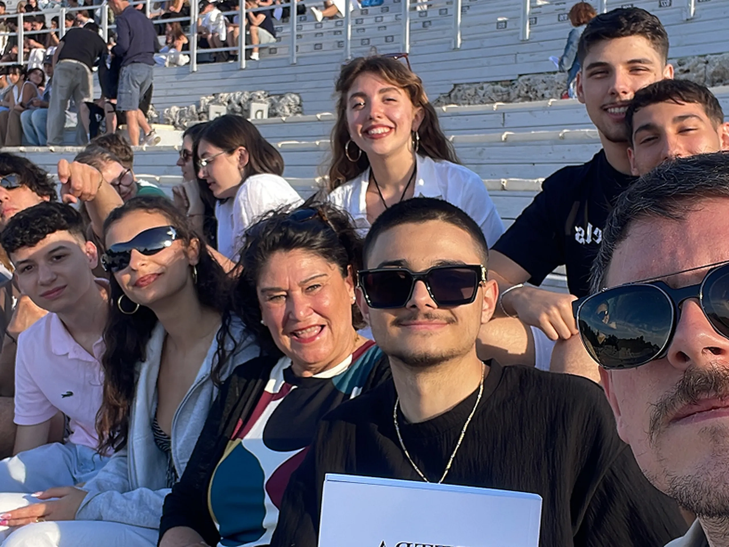 Un gruppo di persone sedute in un anfiteatro all'aperto, che sorridono alla telecamera. Una persona tiene in mano un programma intitolato “Elettra”.