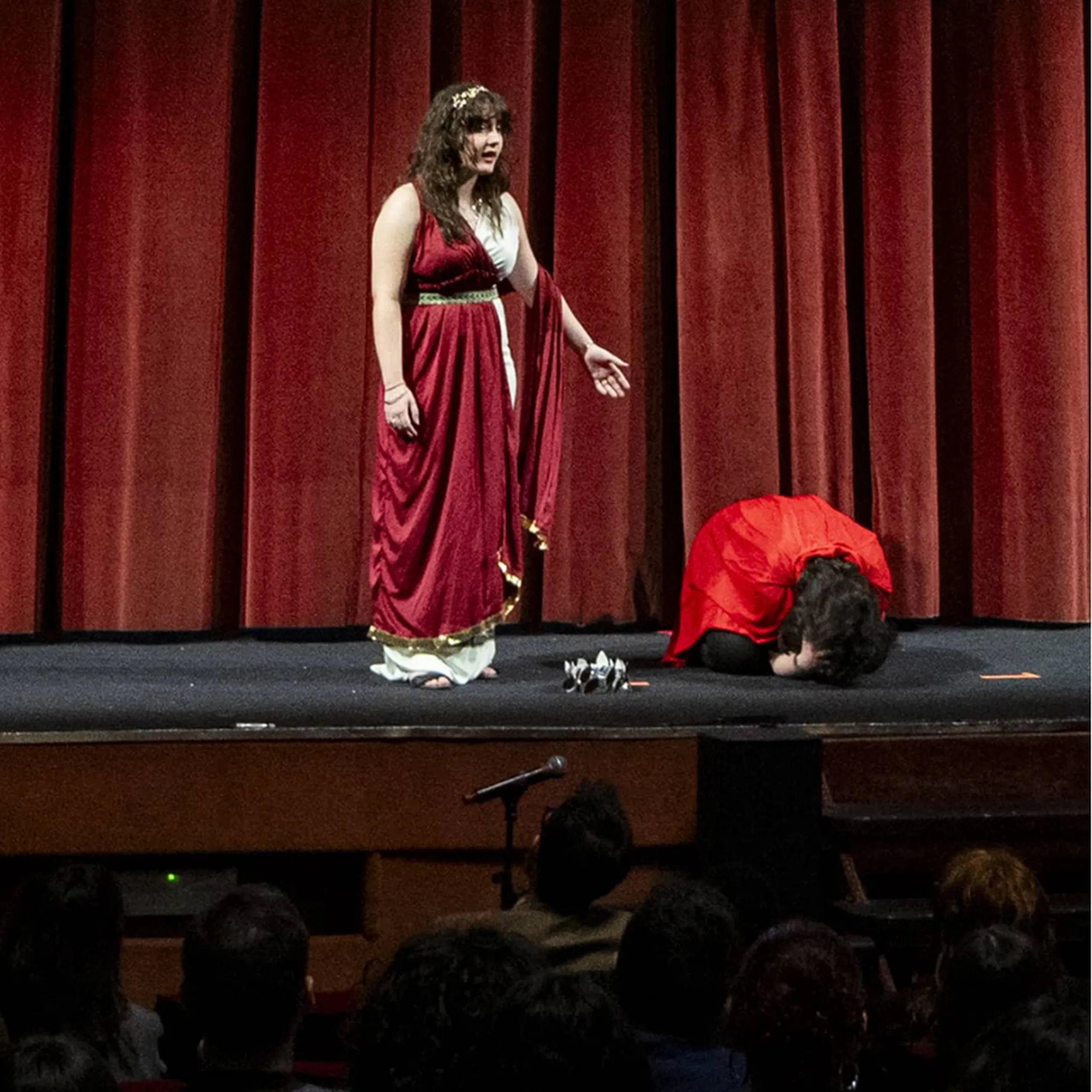 Una donna con un vestito rosso e bianco è in piedi su un palco e gesticola, mentre un'altra persona vestita di rosso è accovacciata accanto a lei. Il pubblico osserva.