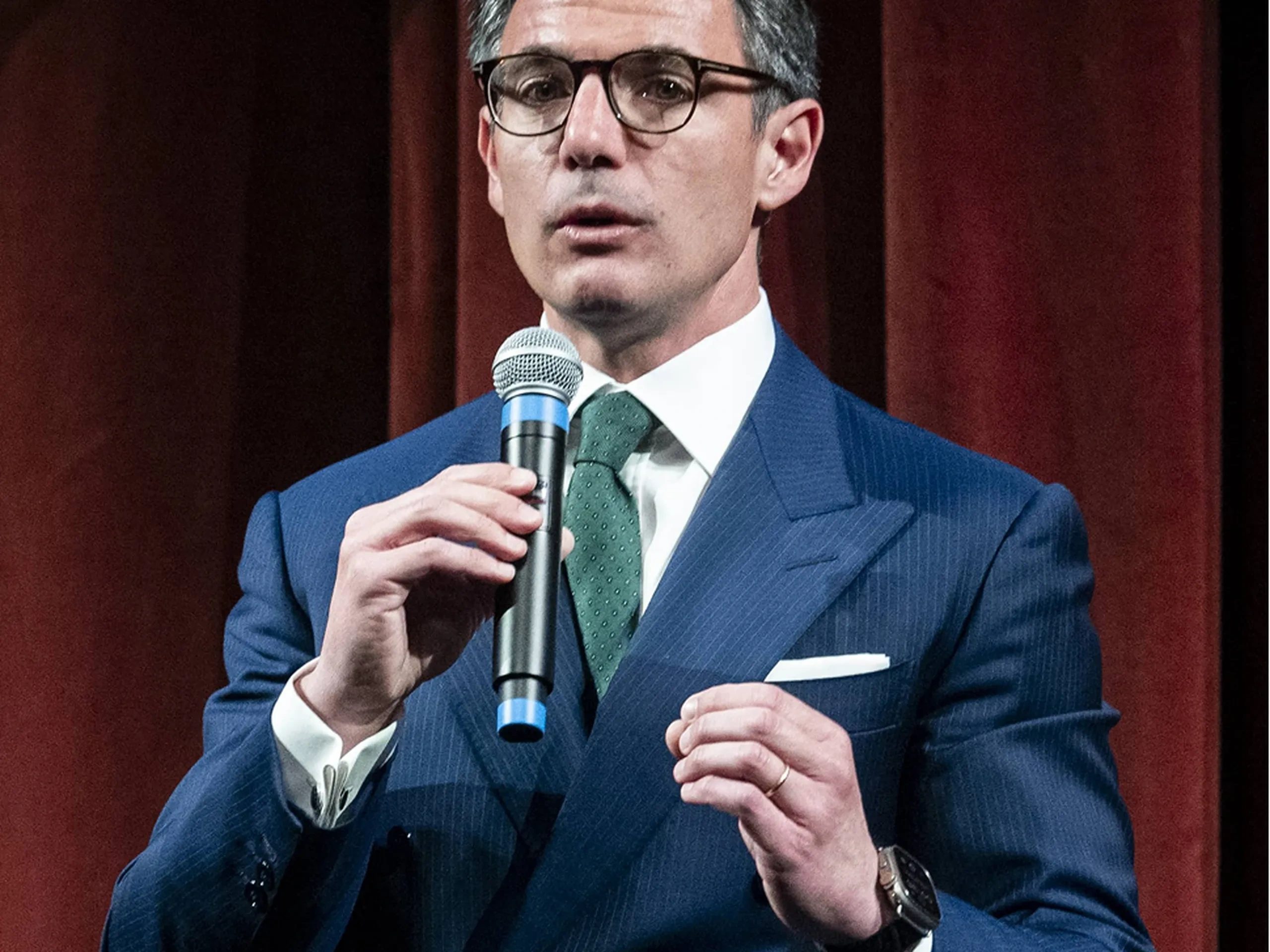Un uomo in abito blu e occhiali sta parlando al microfono davanti a uno sfondo di tende rosso scuro.